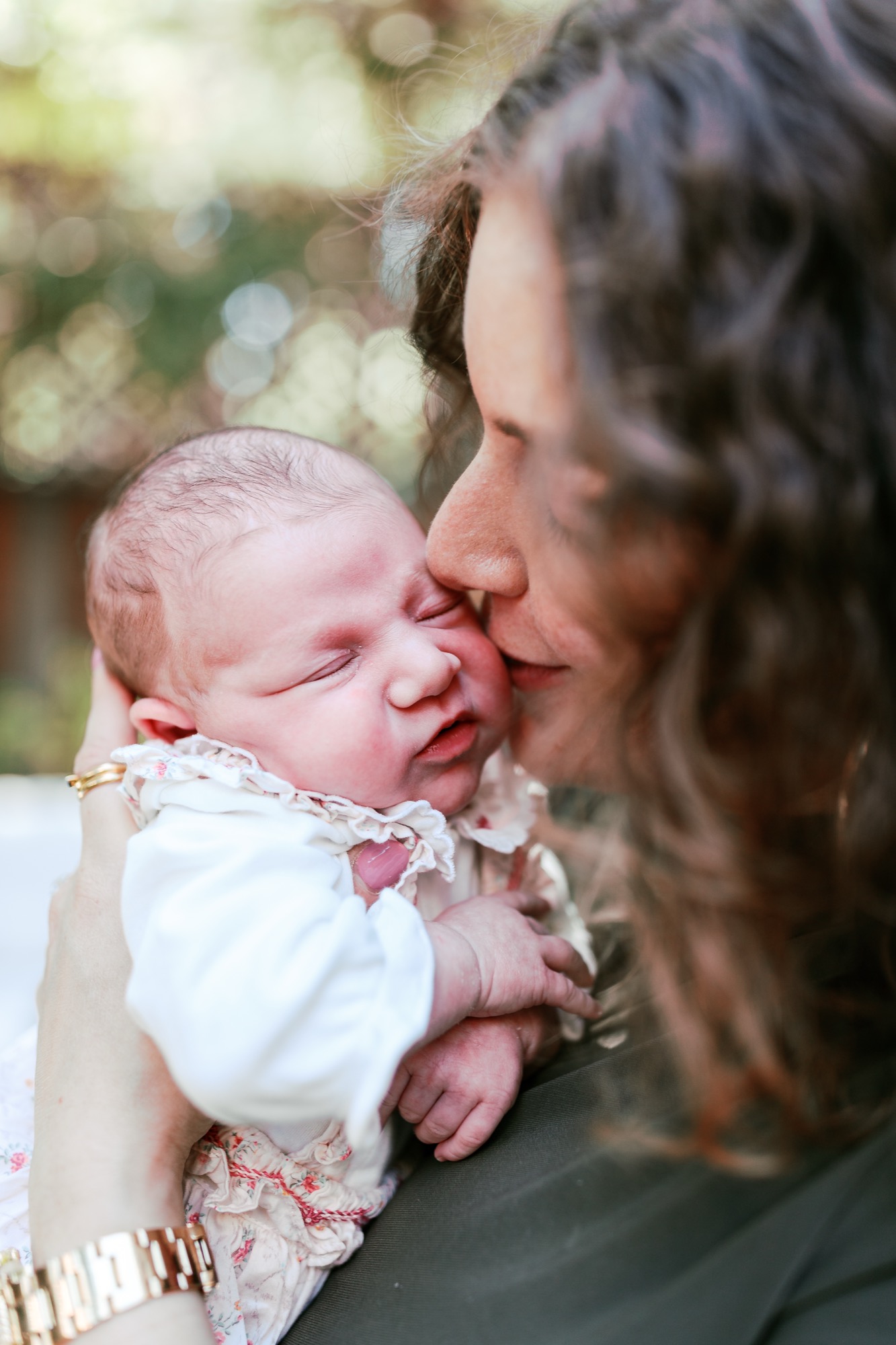 Mother kisses newborn's forehead in soft natural light, baby's eyes closed while wearing white and floral outfit
