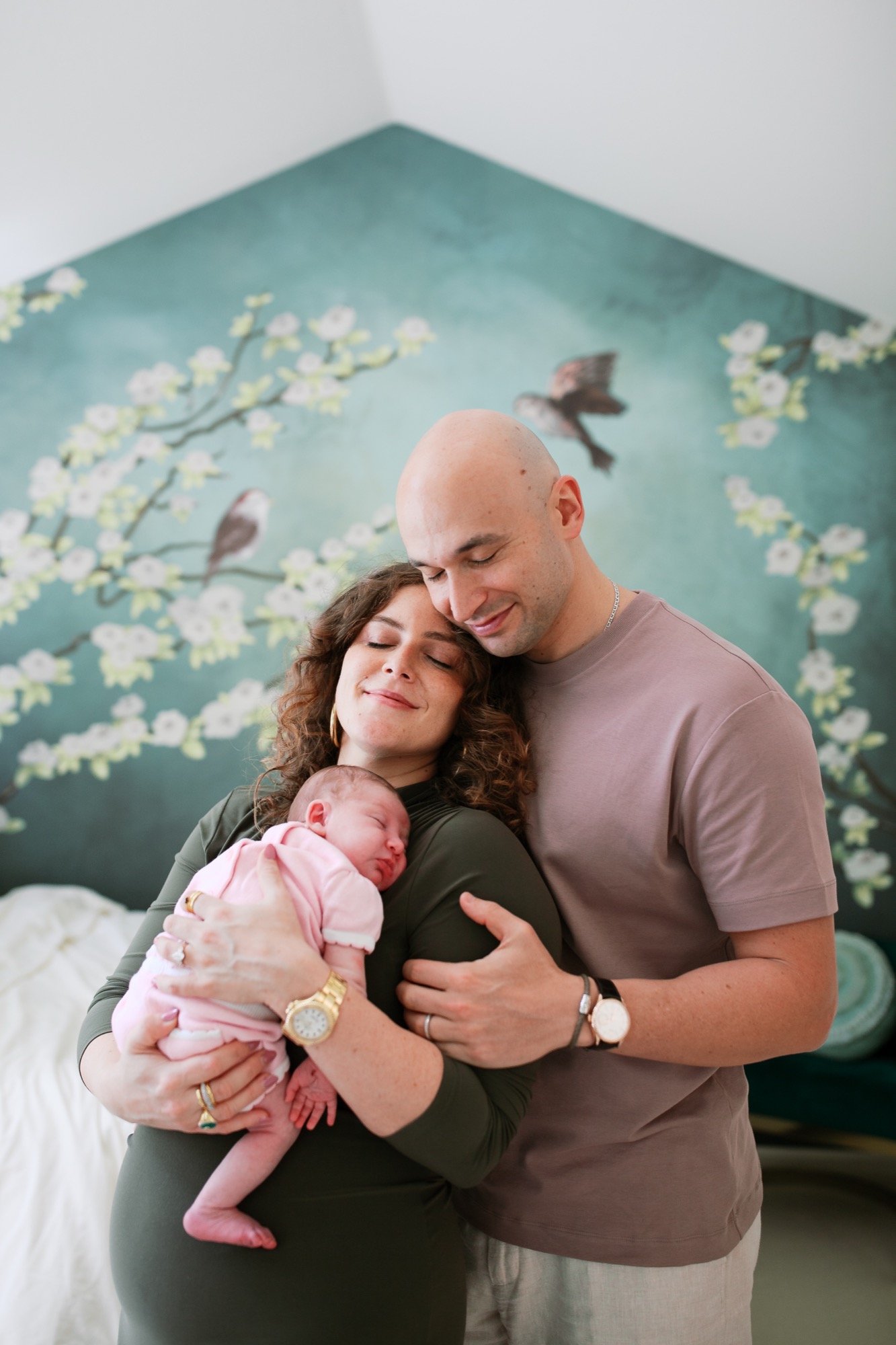 Parents embrace their newborn in pink against a serene teal wall mural of delicate white flowers and perched birds