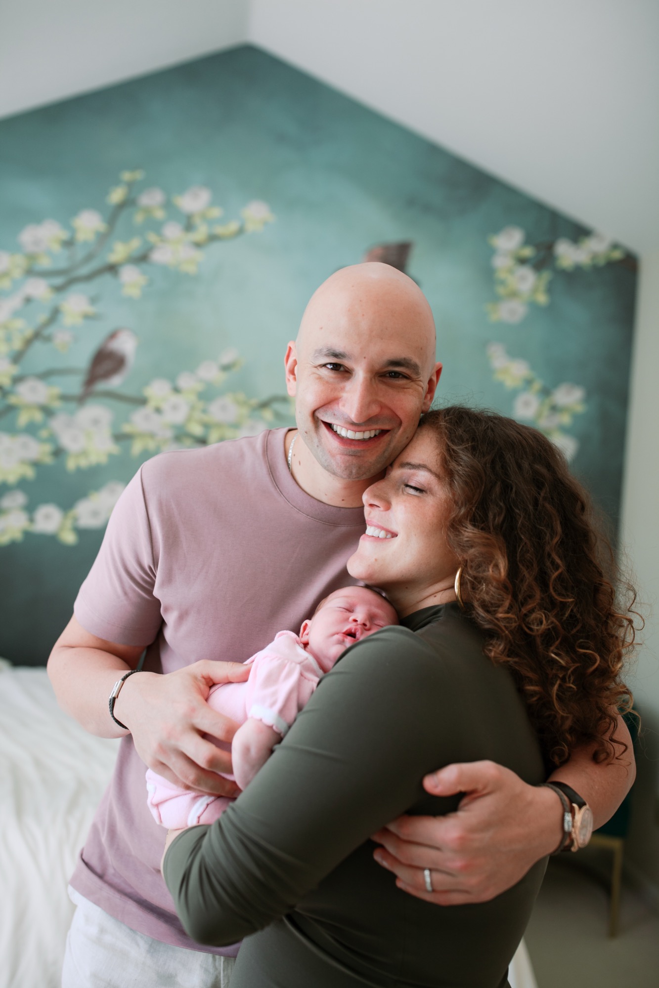 Father beams while mother gazes down at their newborn in pink, bathed in soft window light against a serene sage-green floral backdrop