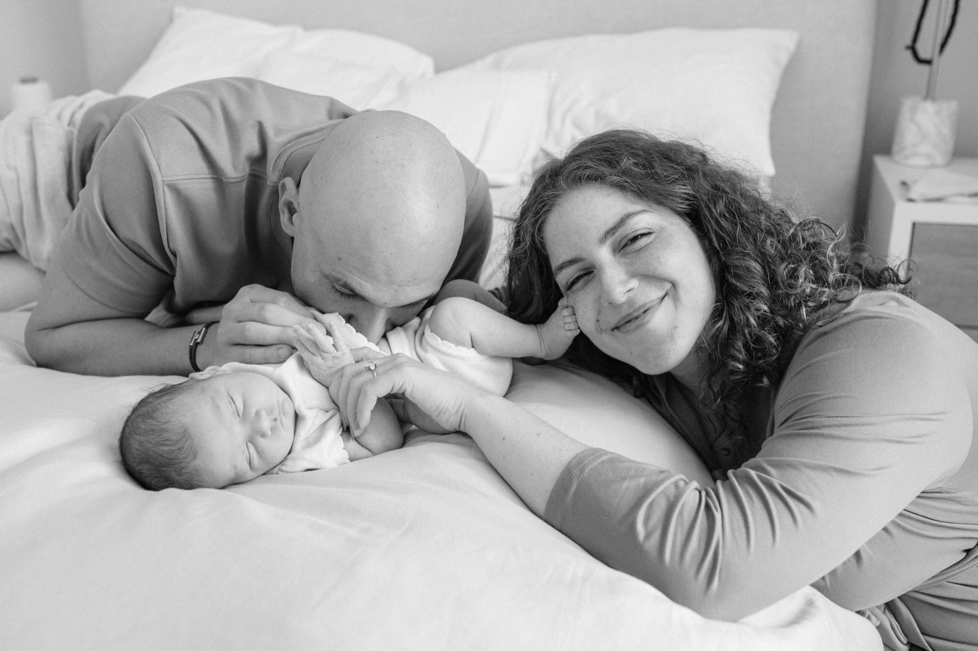 A father kisses his newborn's tiny hands while mom looks on with joy, lying together on the bed (in-home lifestyle newborn photography in Marin County)