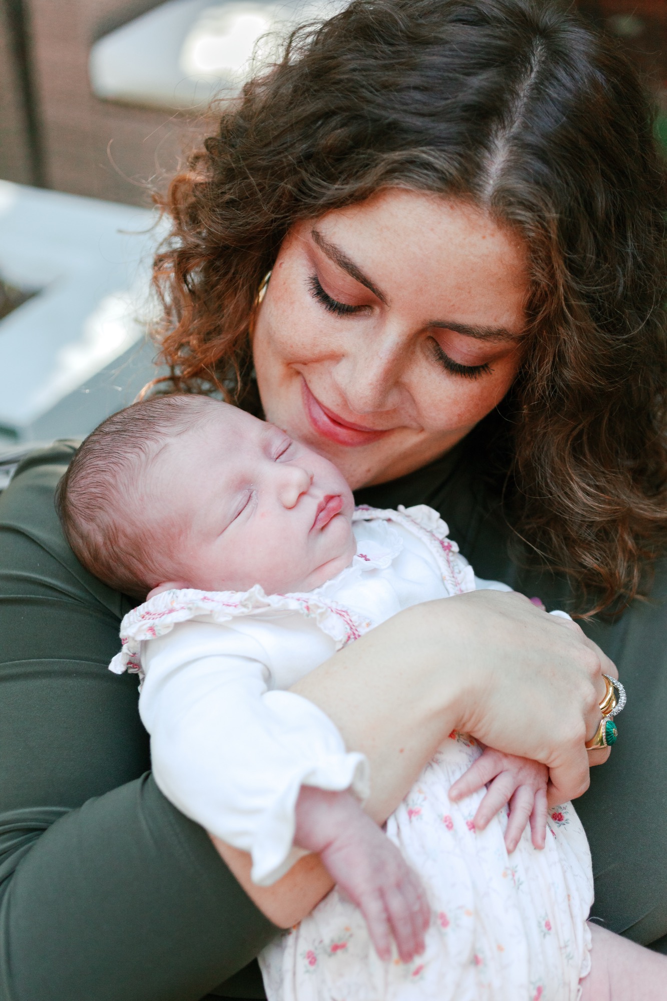 Mother gazes tenderly at her newborn daughter in white floral outfit, natural window light softly illuminating their intimate moment together