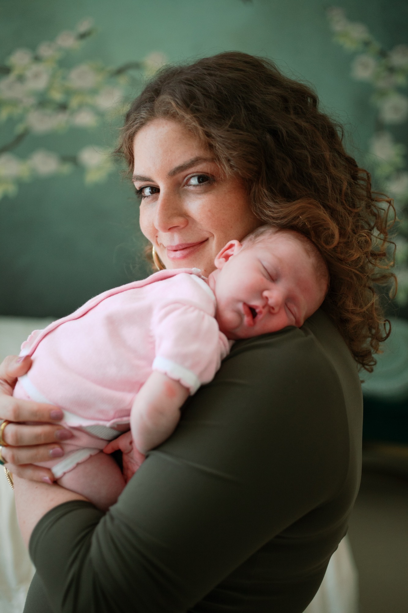 Mother cradles her sleeping newborn against her shoulder, soft natural light catching their connection