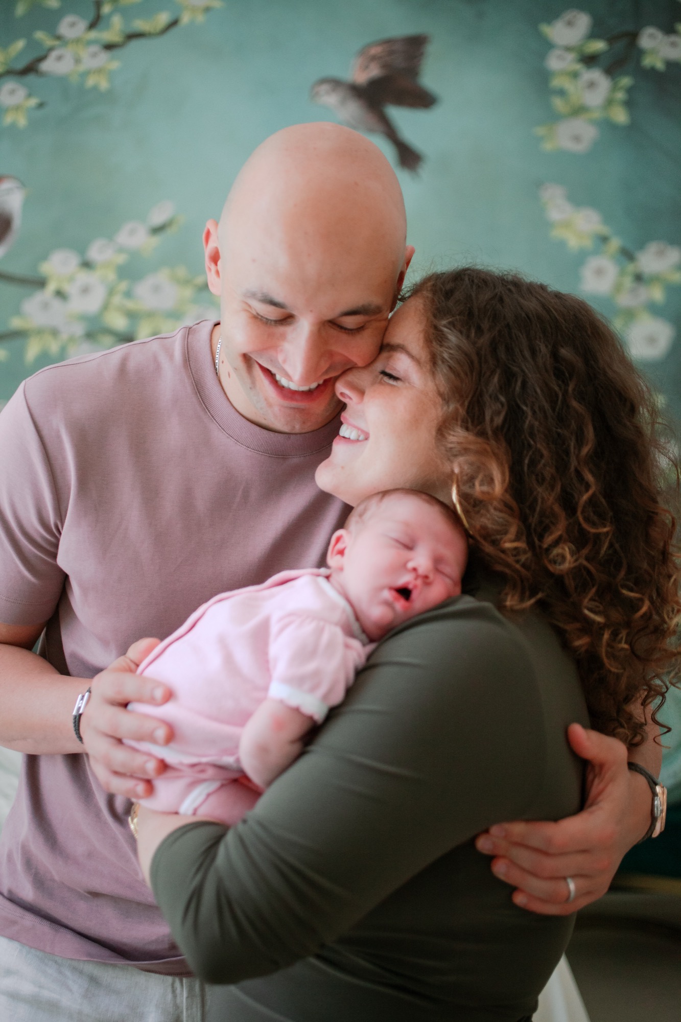 Parents smile warmly while holding their sleeping newborn in pink against a dreamy turquoise floral backdrop with soft natural light