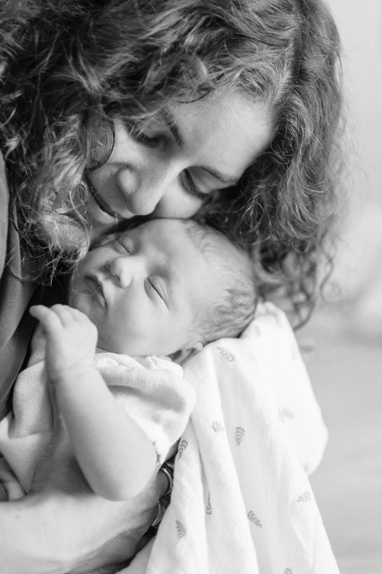 Older sibling gazes tenderly downward at newborn's peaceful face, her curly hair framing both children in soft, diffused light