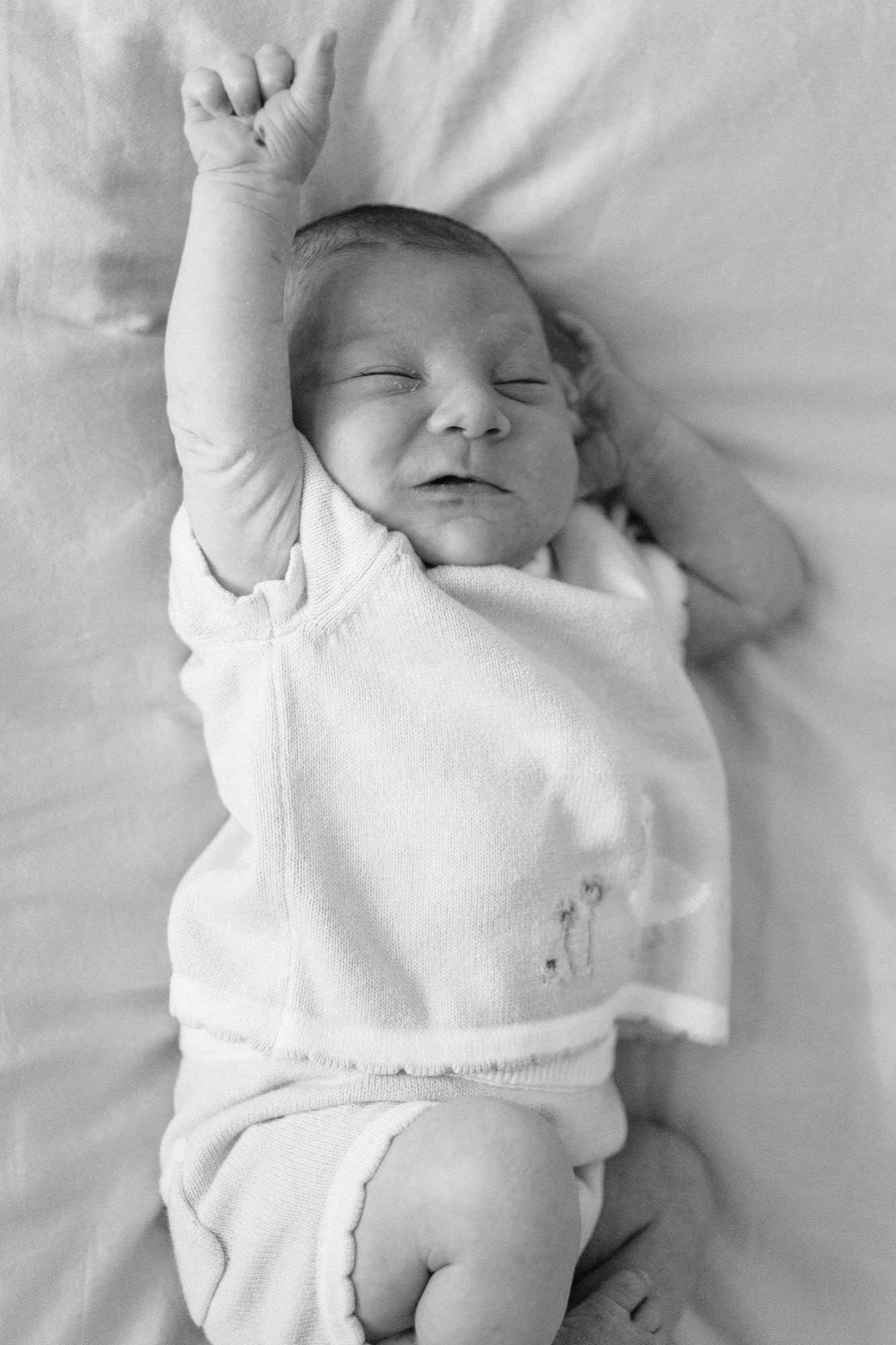 Newborn stretches arm overhead in peaceful sleep, swaddled in soft white cotton against white bedding