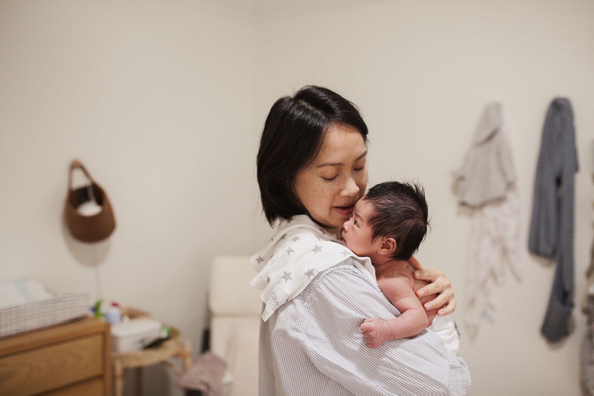 Mother gazes down tenderly at newborn wrapped in white, cradled against her shoulder in soft natural light
