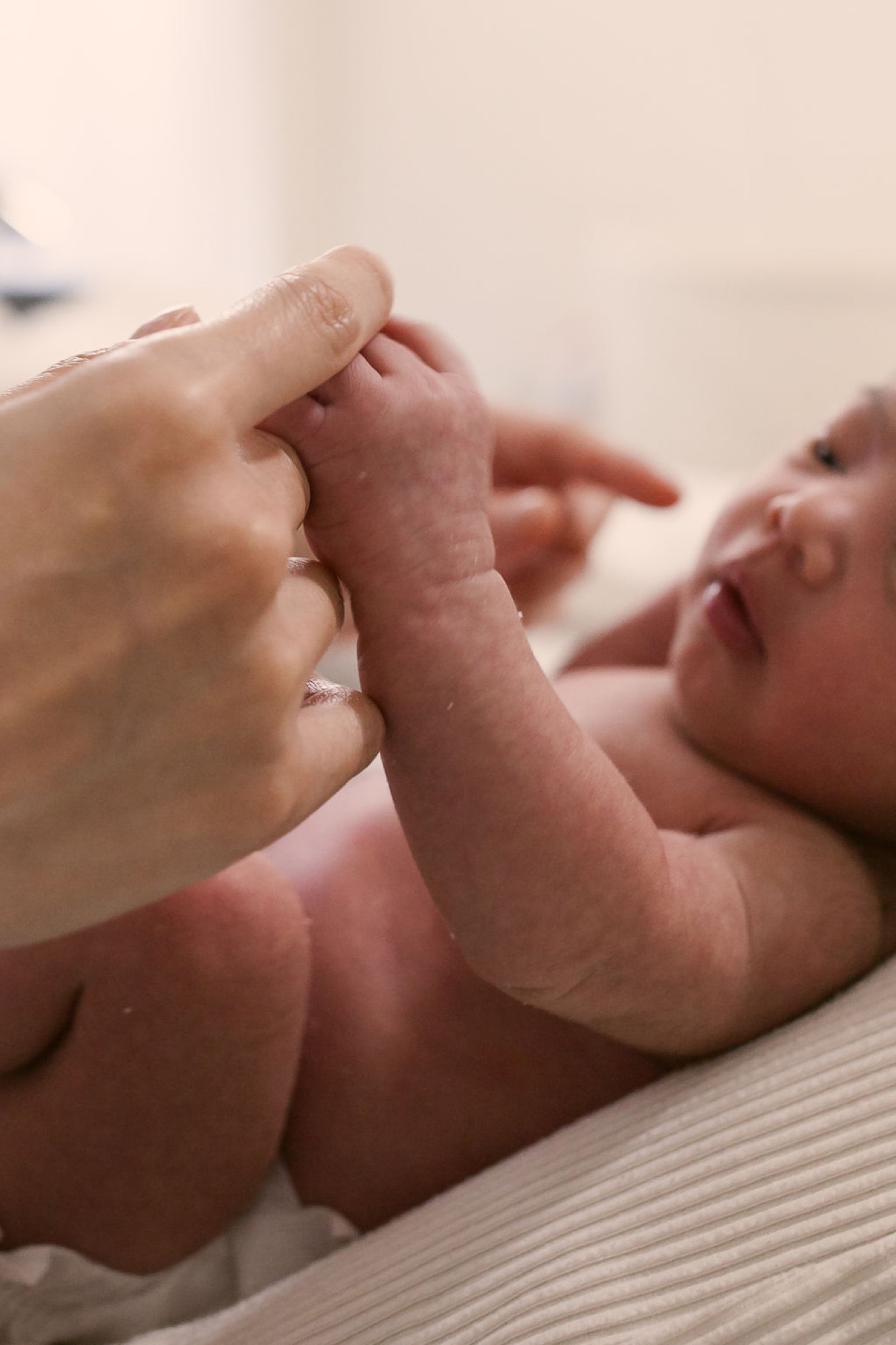 A newborn's tiny hand grasps an adult finger in a tender moment, soft natural light illuminating delicate skin and the intimate bond between caregiver and infant