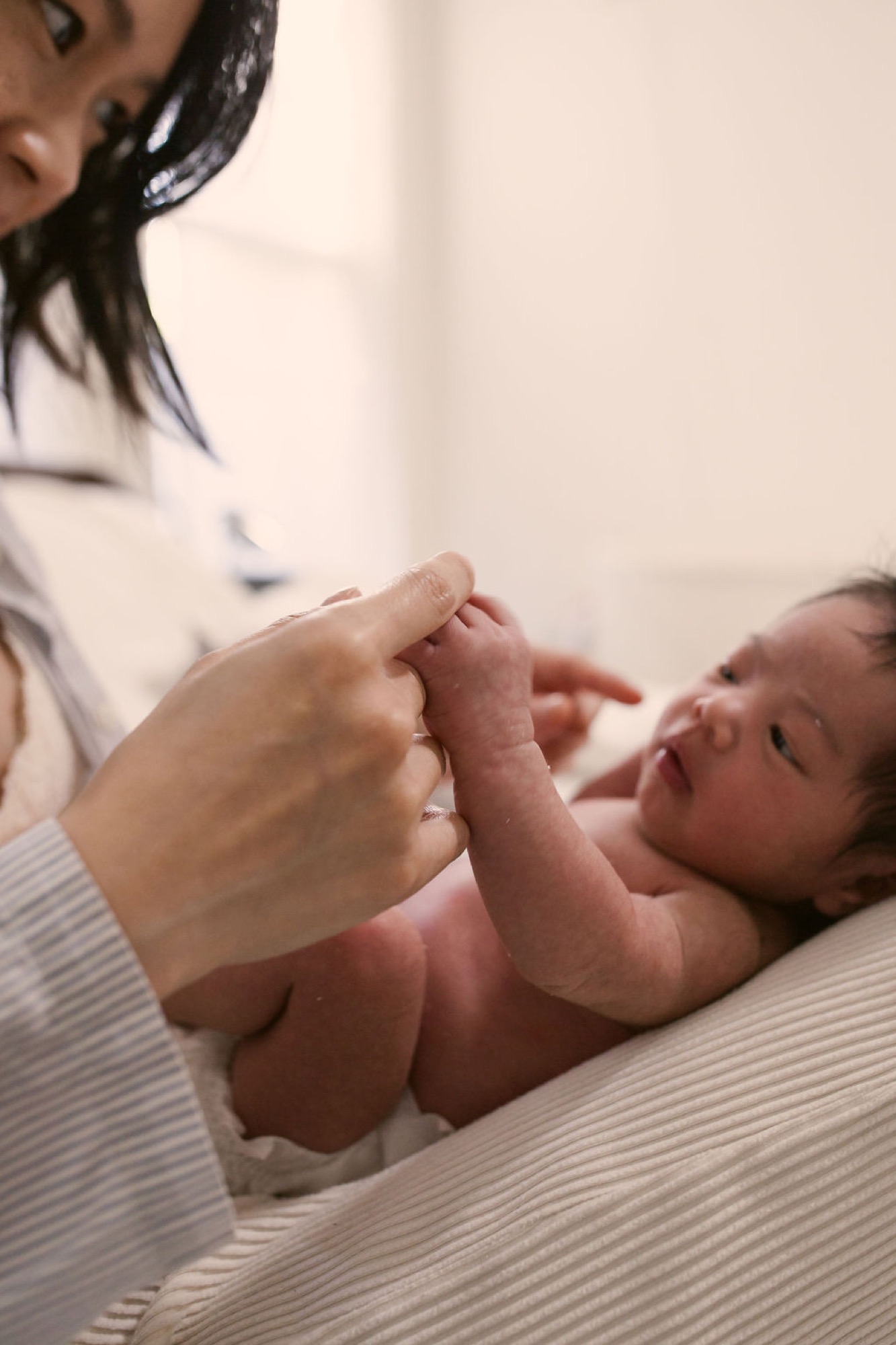 A newborn's tiny hand grasps an adult's finger in a tender moment, wrapped in cream-colored fabric during an intimate indoor session