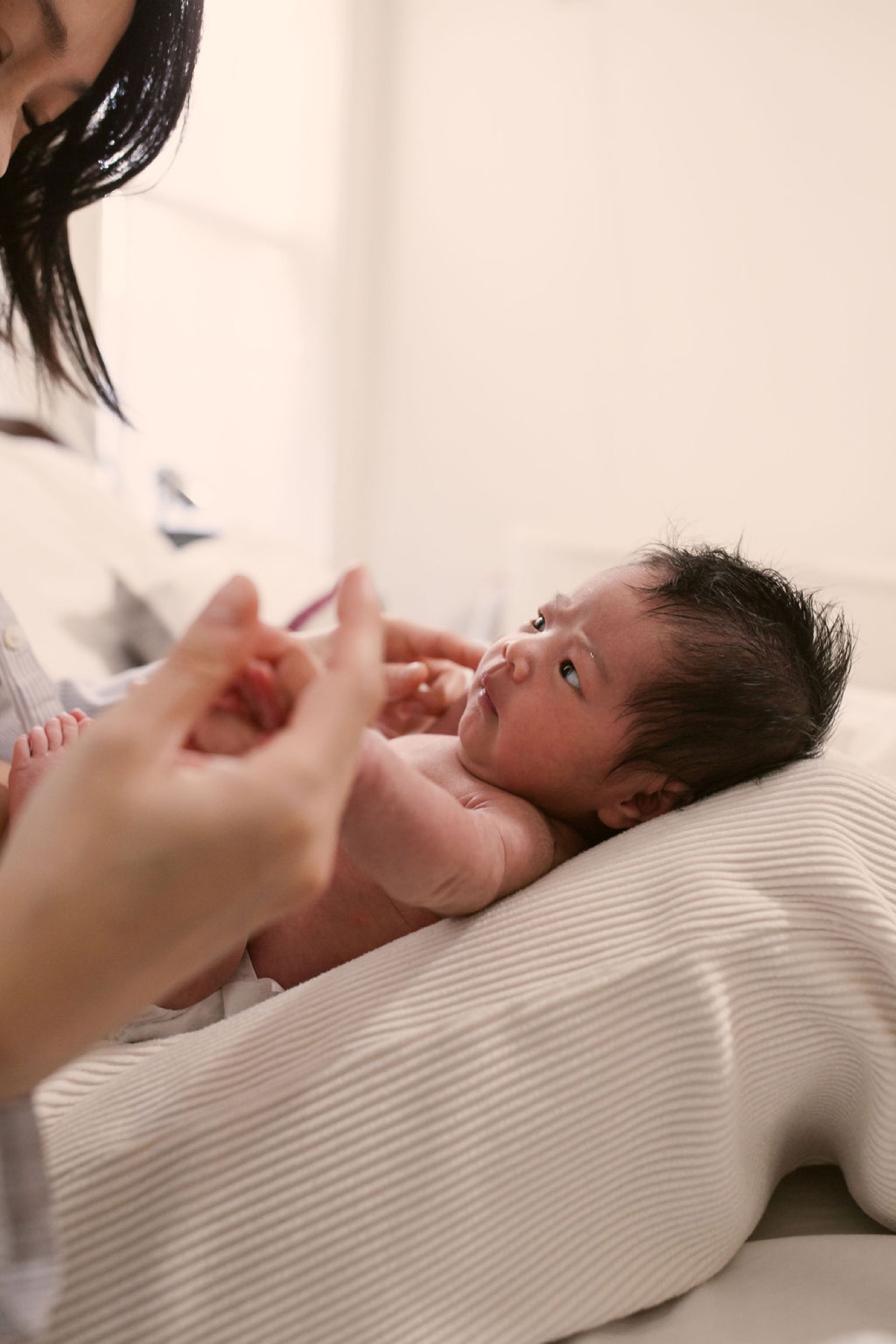 Newborn gazes upward while mother leans in close, their hands gently touching on soft cream bedding in warm natural light