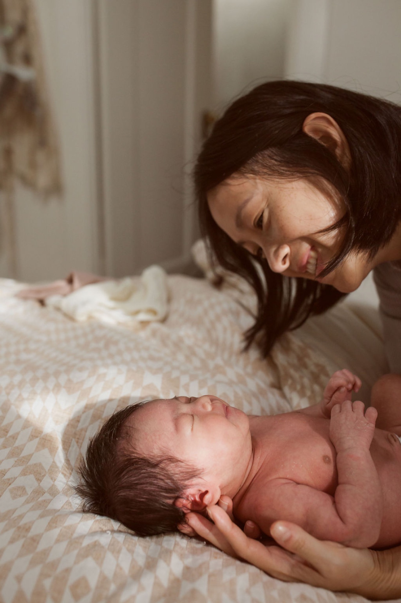 An older sister gazes down tenderly at her sleeping newborn brother on a cream textured blanket in soft natural light