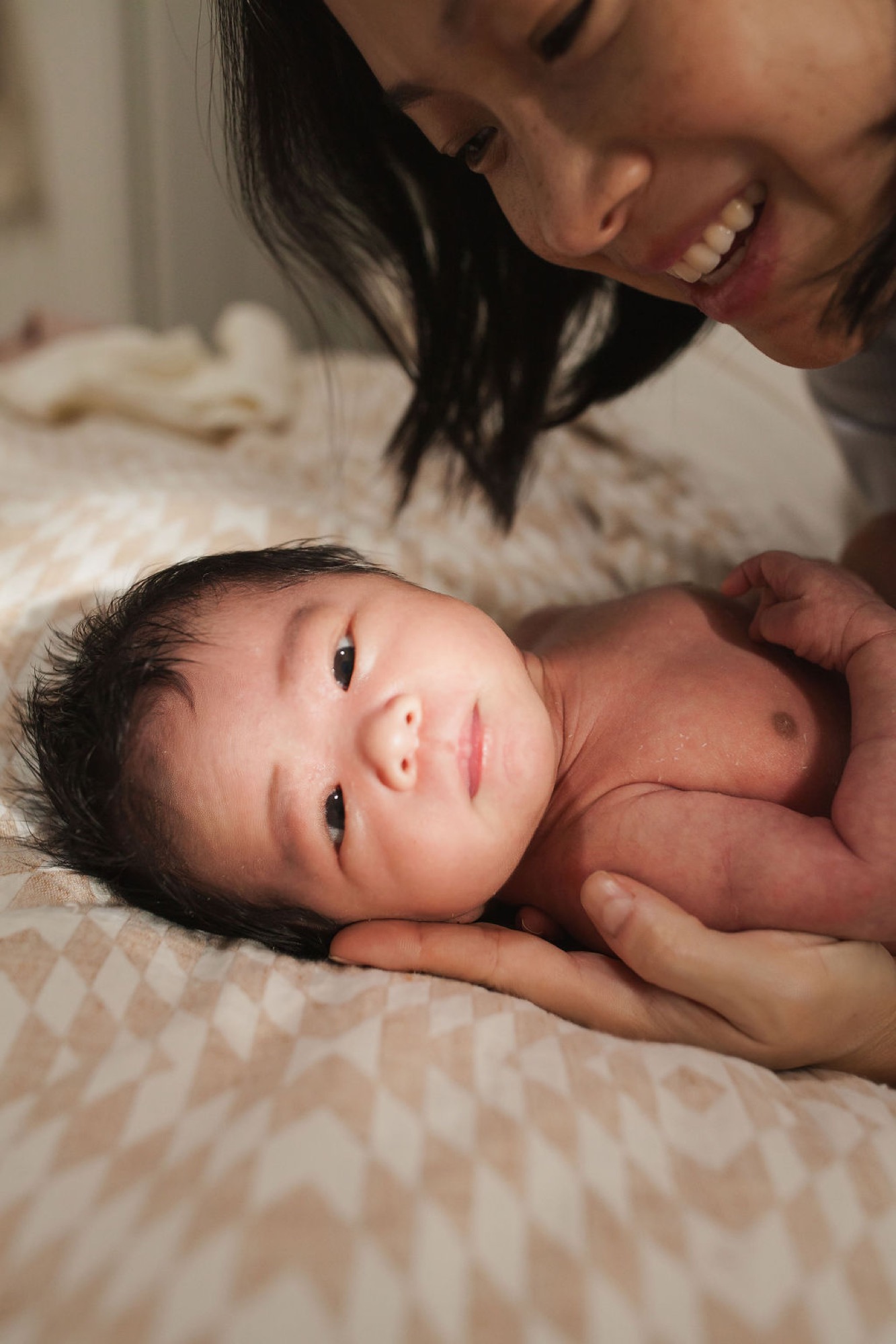 A mother beams down at her newborn, who gazes ahead with alert eyes while nestled in her supportive hands on cream bedding