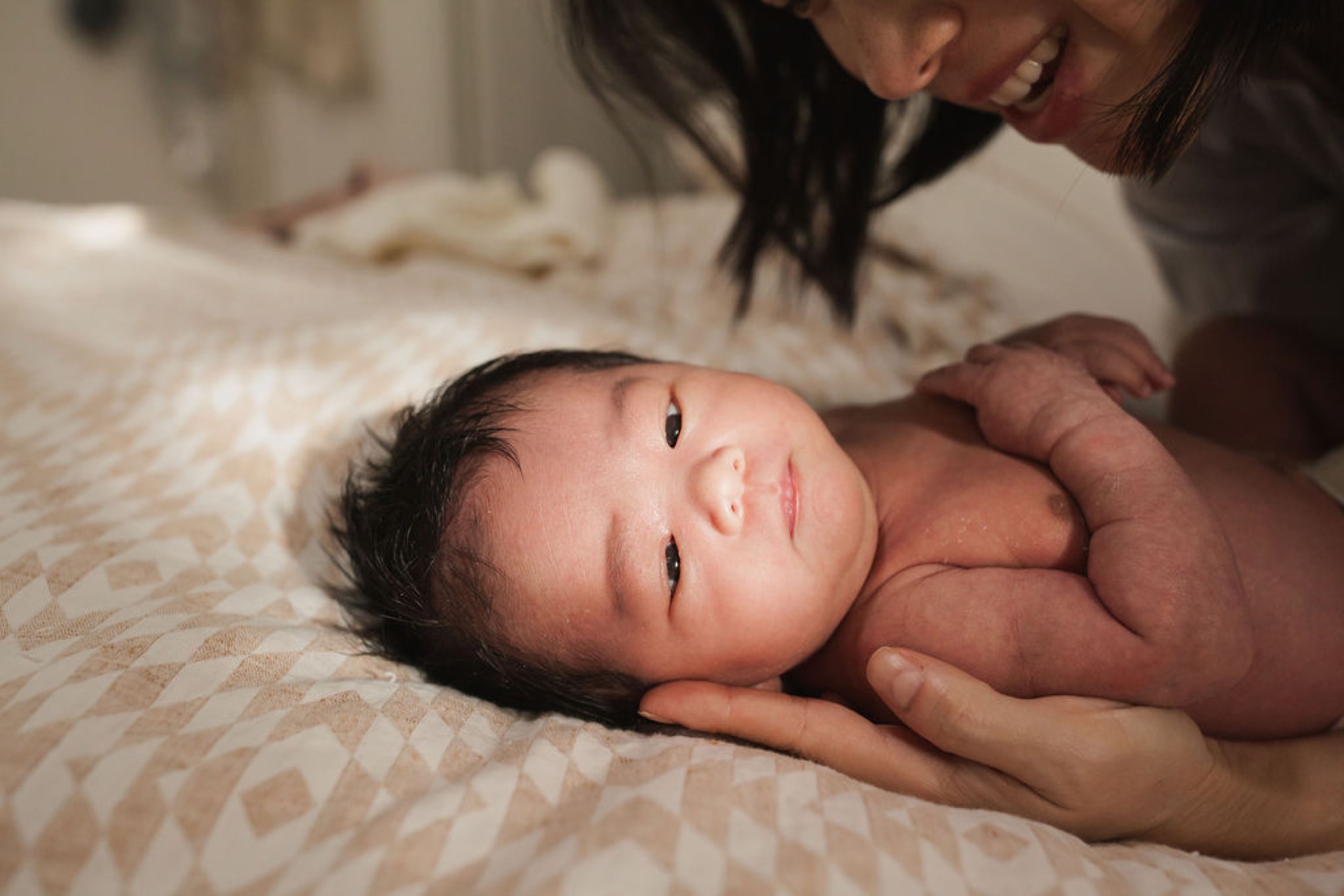 Newborn with dark hair lies on a cream patterned blanket, eyes open and alert, cradled by a parent's hands from above