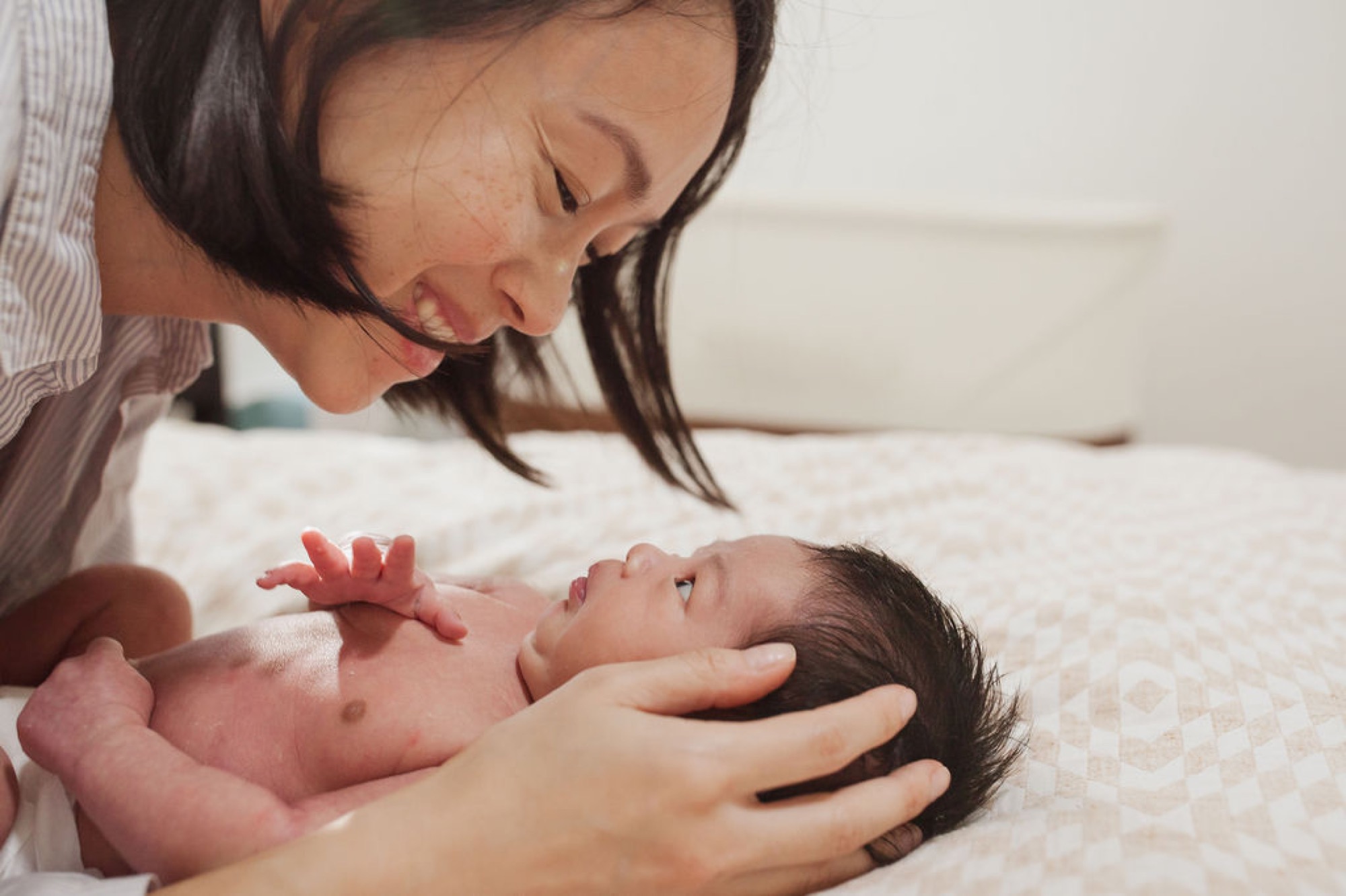 Mother gazes tenderly at newborn while cradling infant's head, soft natural light illuminating their intimate bonding moment on white bedding