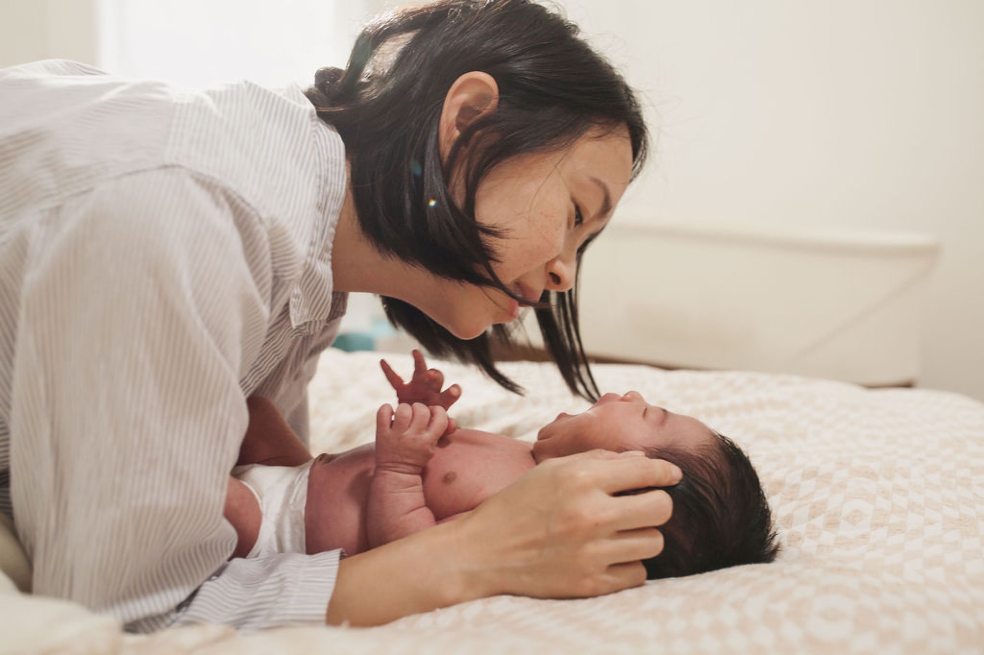 Mother gazes tenderly at newborn while cradling infant on white bedding, soft natural light illuminating intimate bonding moment