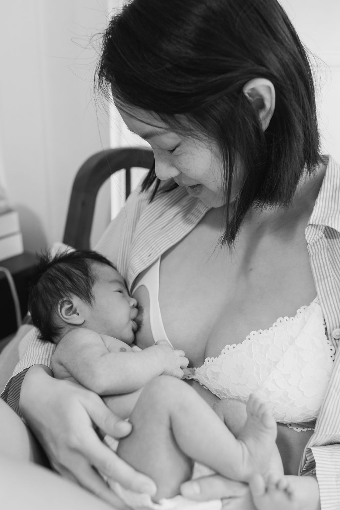 Mother gazes down tenderly at nursing newborn in soft window light, intimate black and white portrait capturing the quiet bond between them