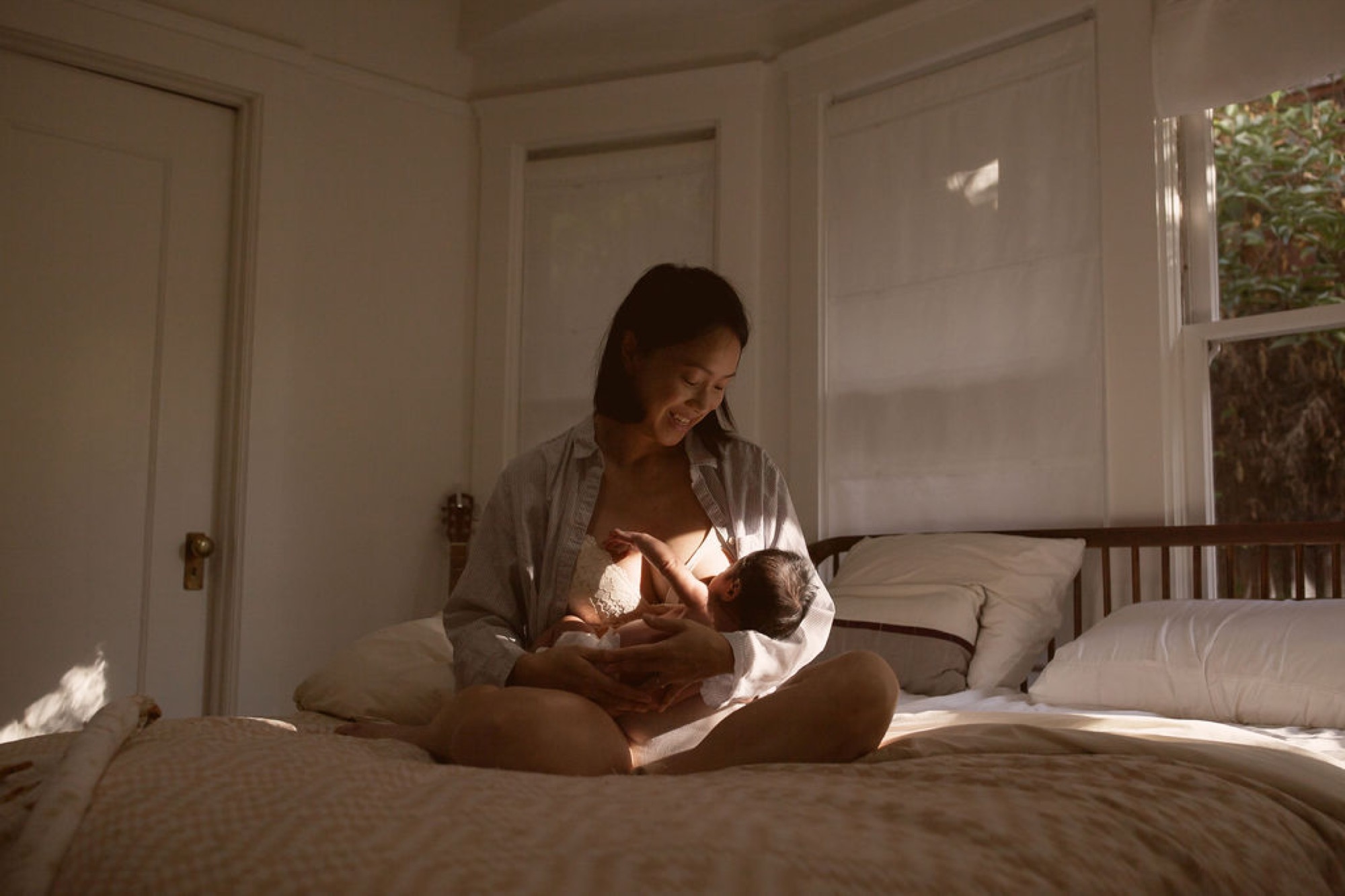 A mother sits cross-legged on a sunlit bed, gazing down at her newborn with a gentle smile as warm light streams through the window