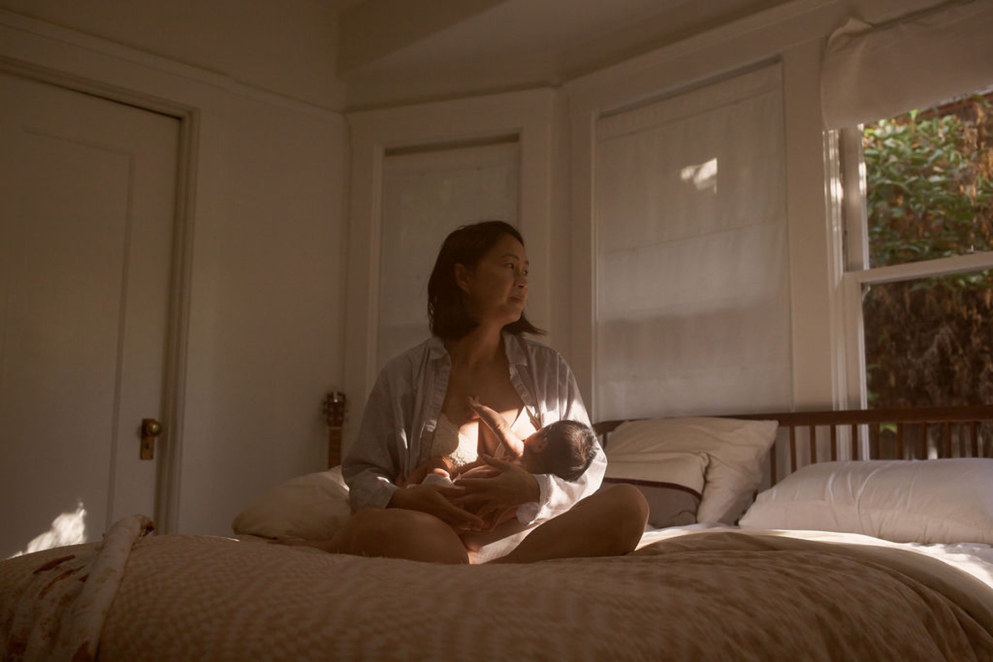 Mother gazes thoughtfully out the window while cradling her newborn in golden afternoon light flooding the serene bedroom