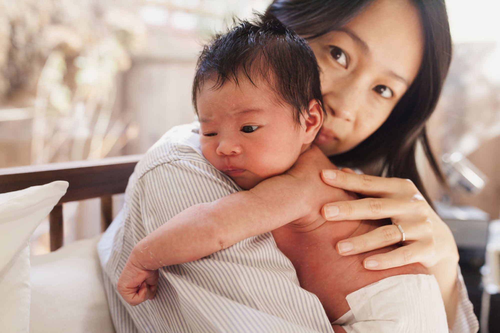Mother gazes at camera while cradling her newborn, who looks downward in a soft striped outfit against a neutral home interior