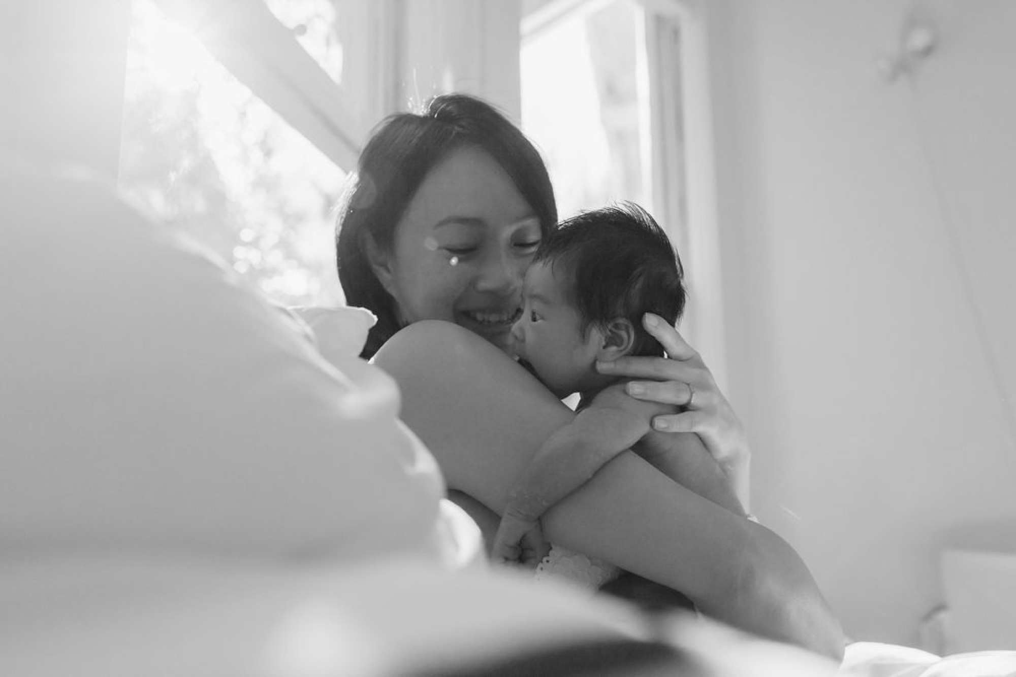 Mother and infant share an intimate moment by bright window light, their foreheads nearly touching as both beam with genuine joy and connection