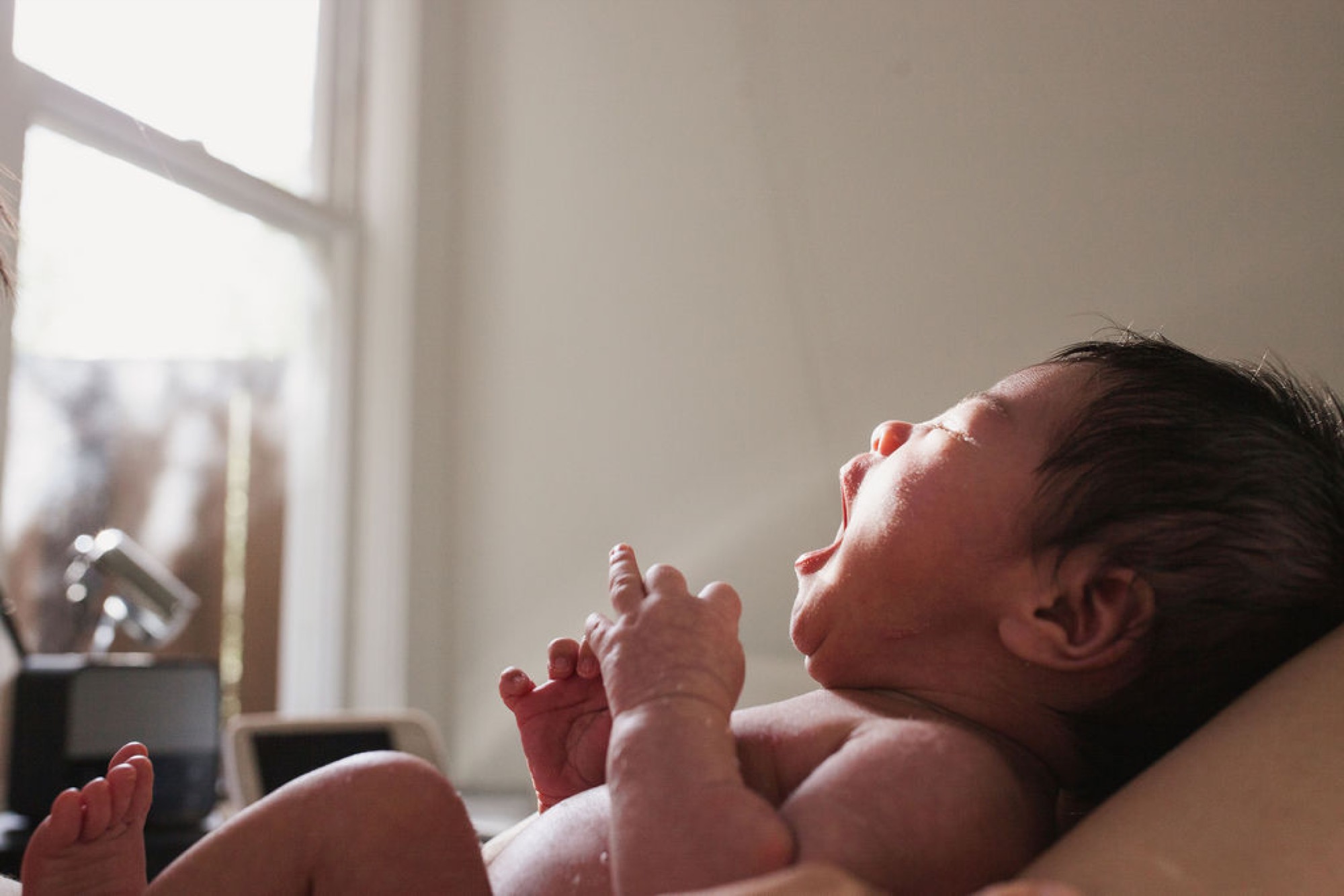 Newborn stretches tiny hands toward bright window light, head tilted back in peaceful contentment during golden afternoon