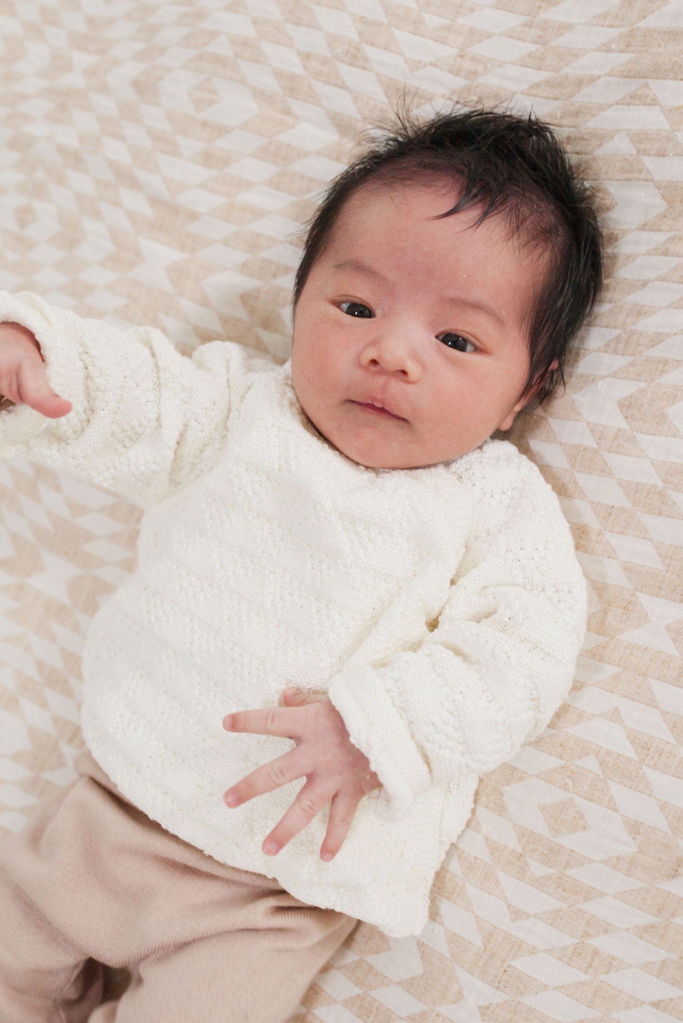Newborn wrapped in cream knit lies on patterned bedding, gazing directly at camera with alert, contemplative expression and relaxed hands