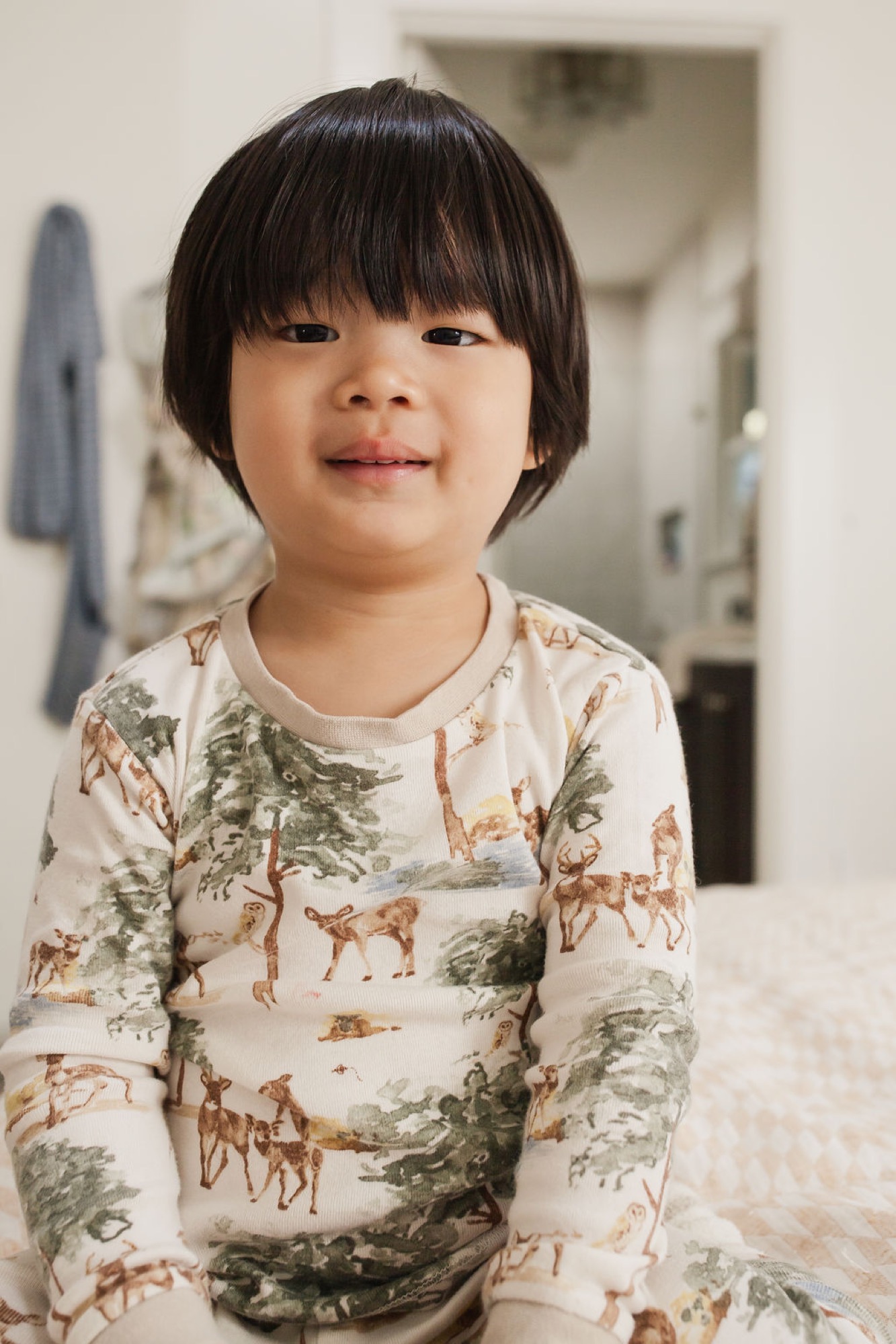 Toddler in woodland-print pajamas smiles gently at camera in bright home interior with natural window light