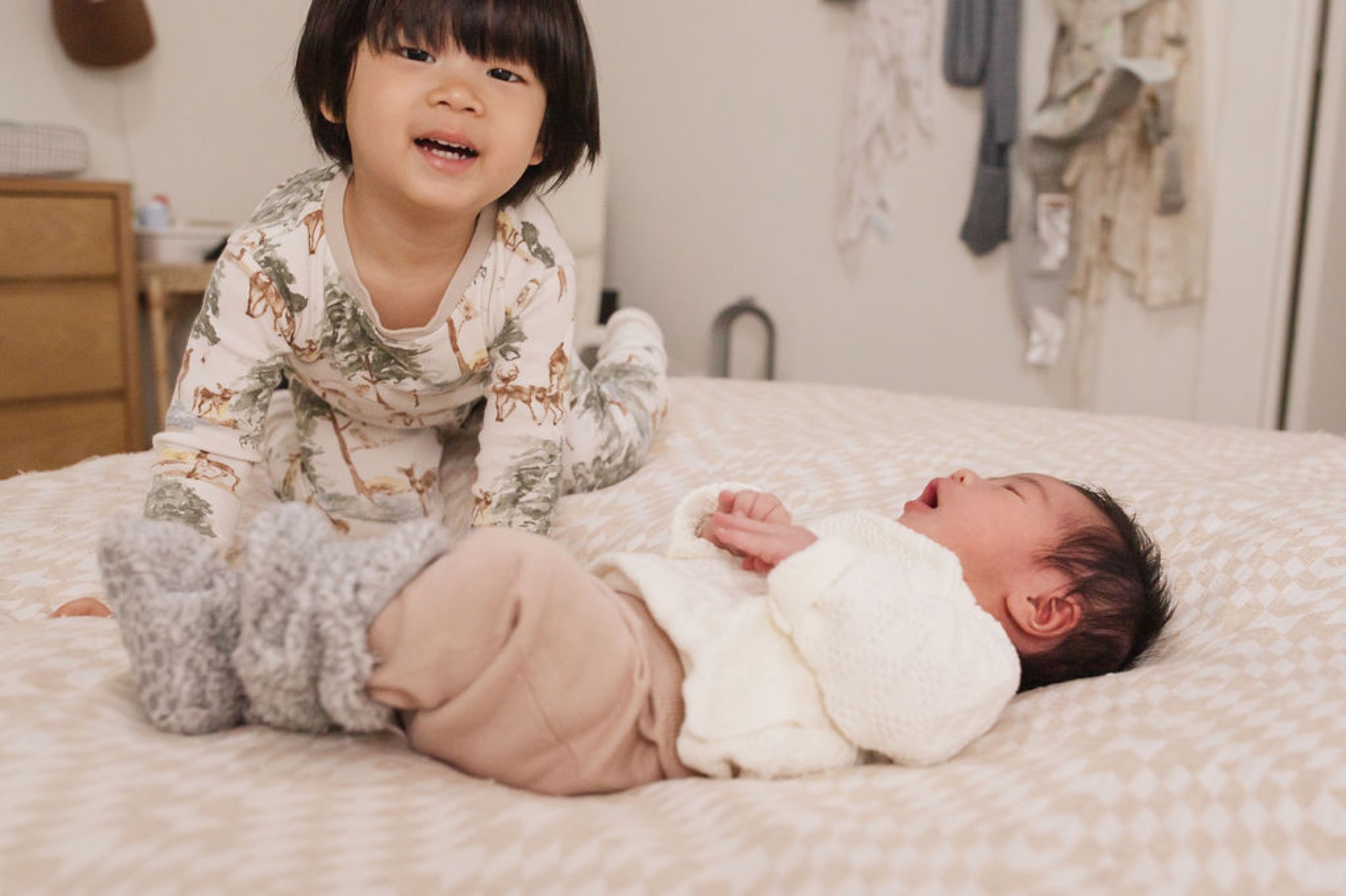 Toddler in floral pajamas sits beside newborn on cream bedding, smiling down at swaddled infant in bright bedroom