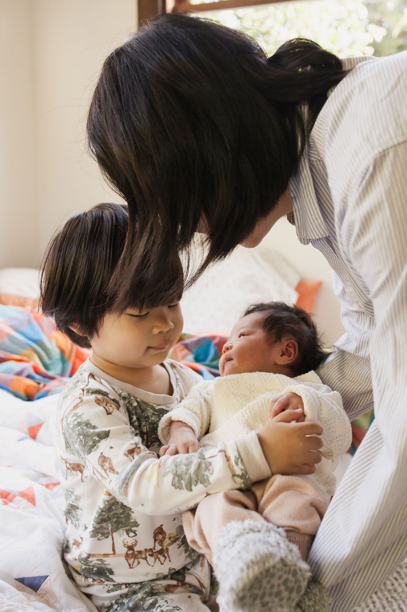 Older sibling in forest-print pajamas gently holds newborn while mother leans in, soft natural light streaming across the bed