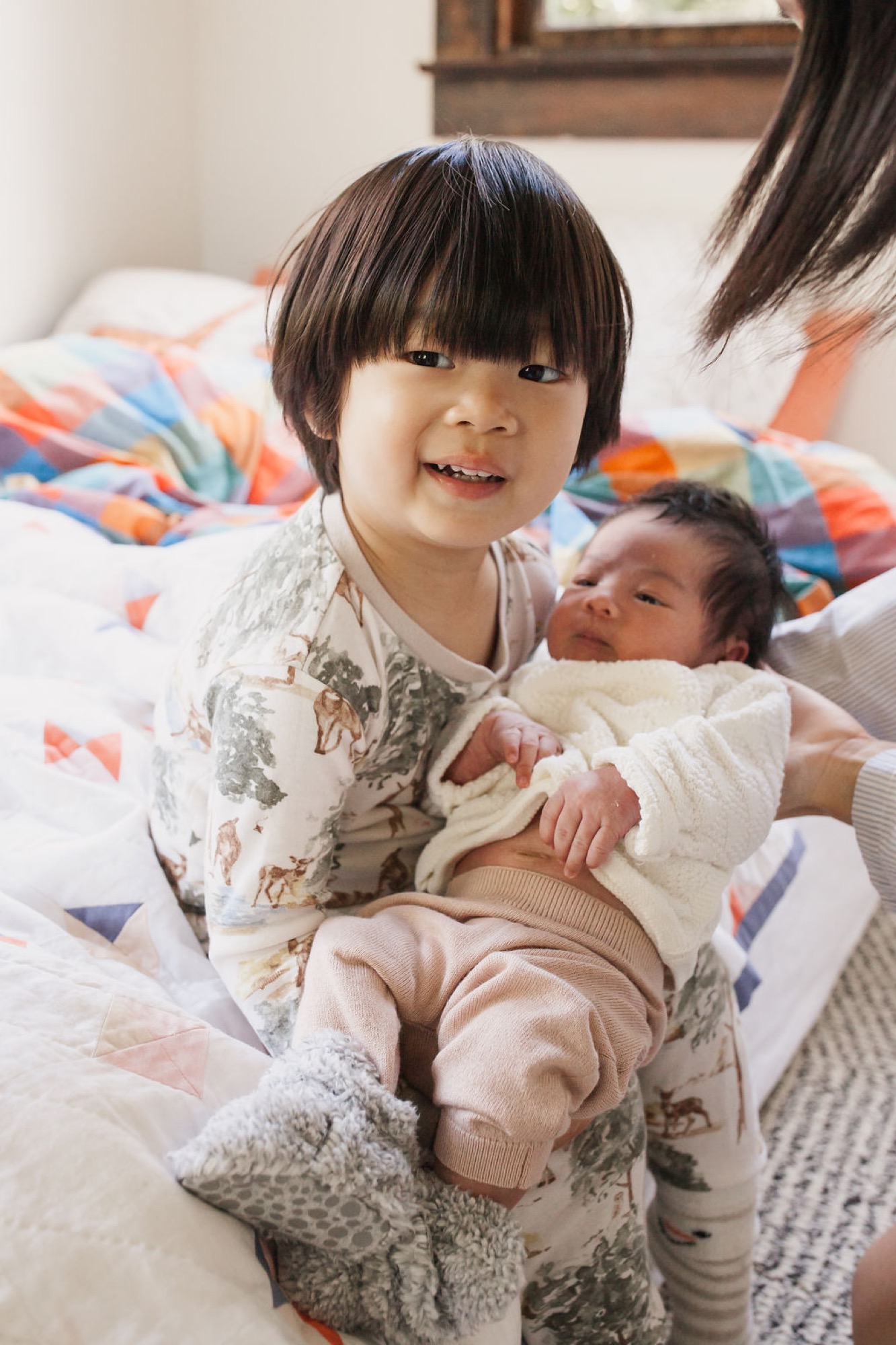 Older sister beams with joy while cradling her newborn sibling on a colorful bed in soft natural light