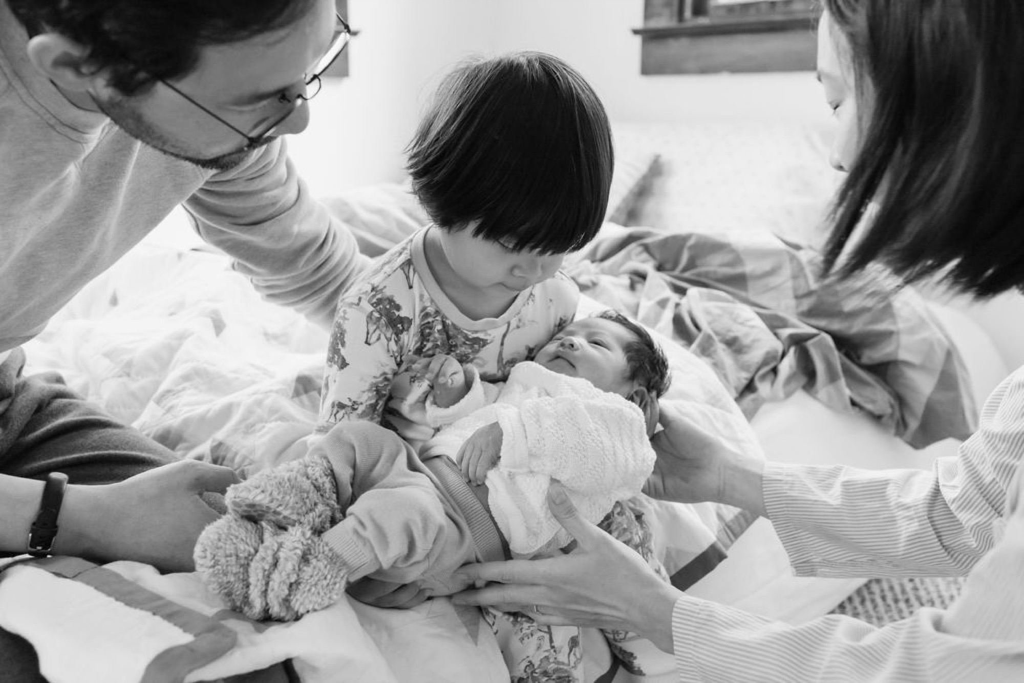 A newborn nestled in her older sister's lap while three adults lean in tenderly, their hands gently touching the infant in soft natural light