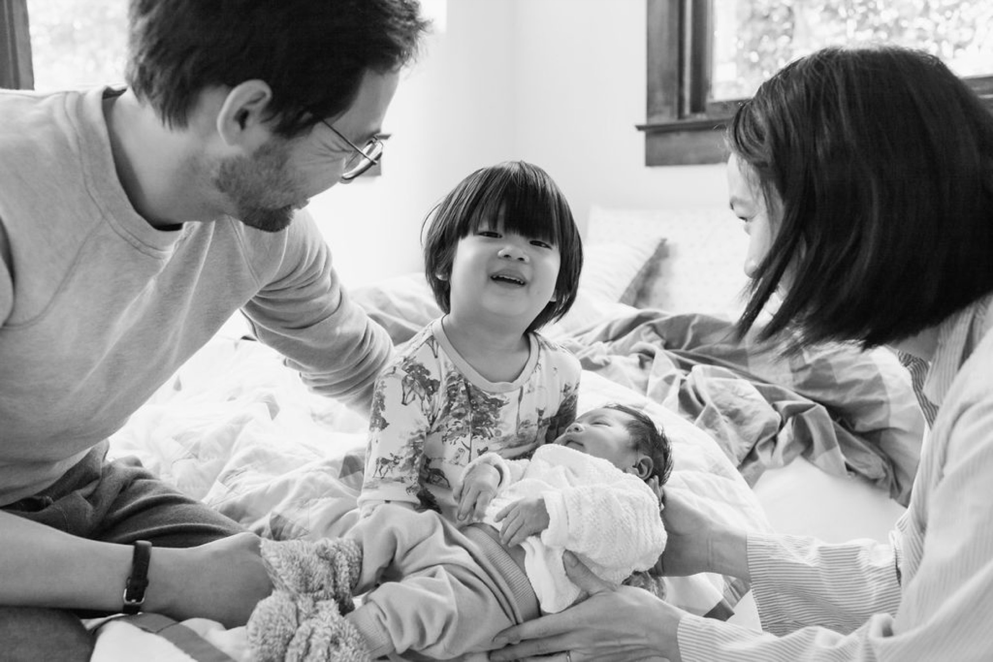 A father in glasses leans toward his beaming toddler as the mother holds their newborn, all gathered on white bedding in soft natural light