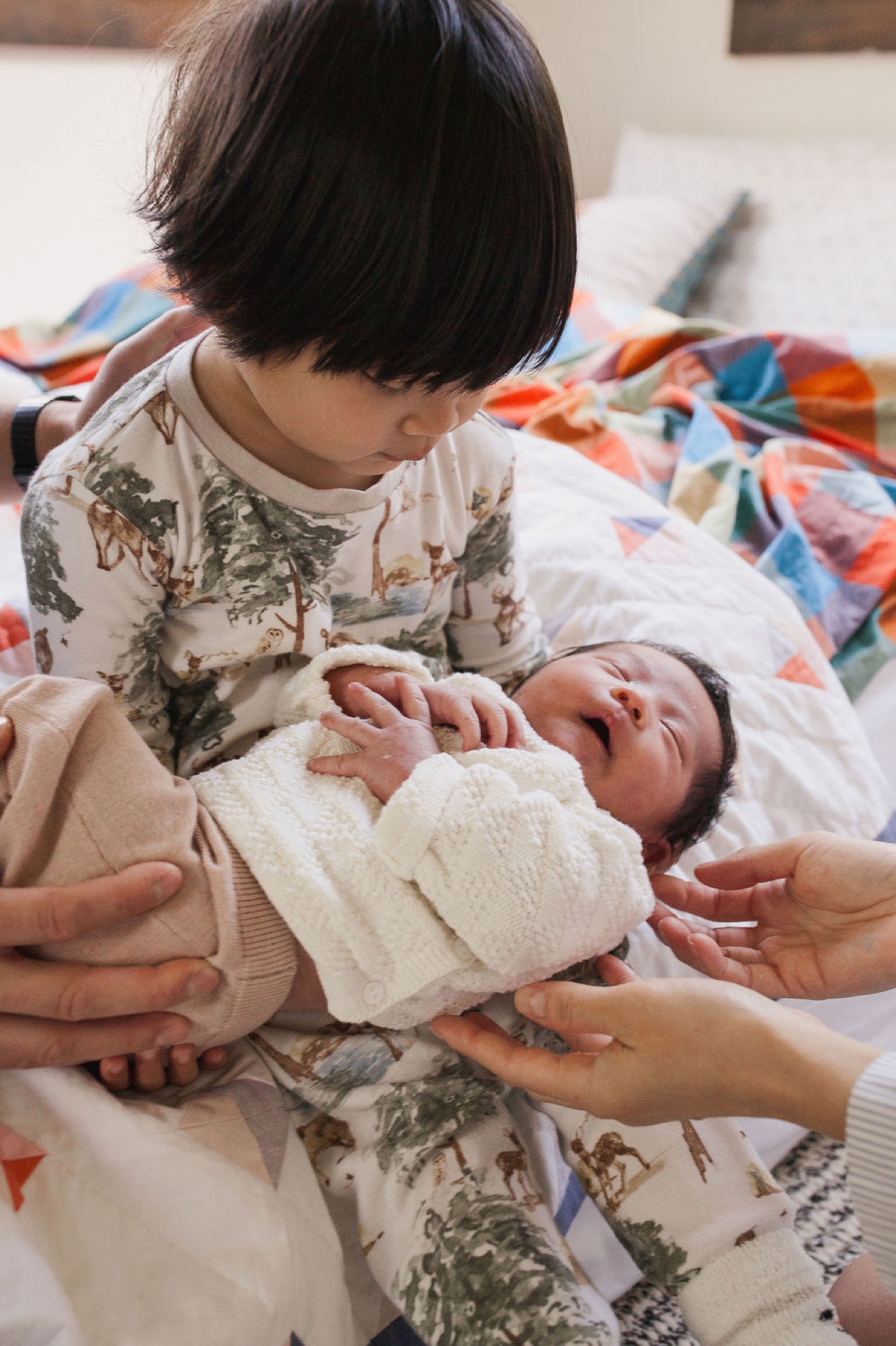 An older sibling gently touches a newborn swaddled in cream fabric while an adult's hands guide the tender moment on a colorful quilt
