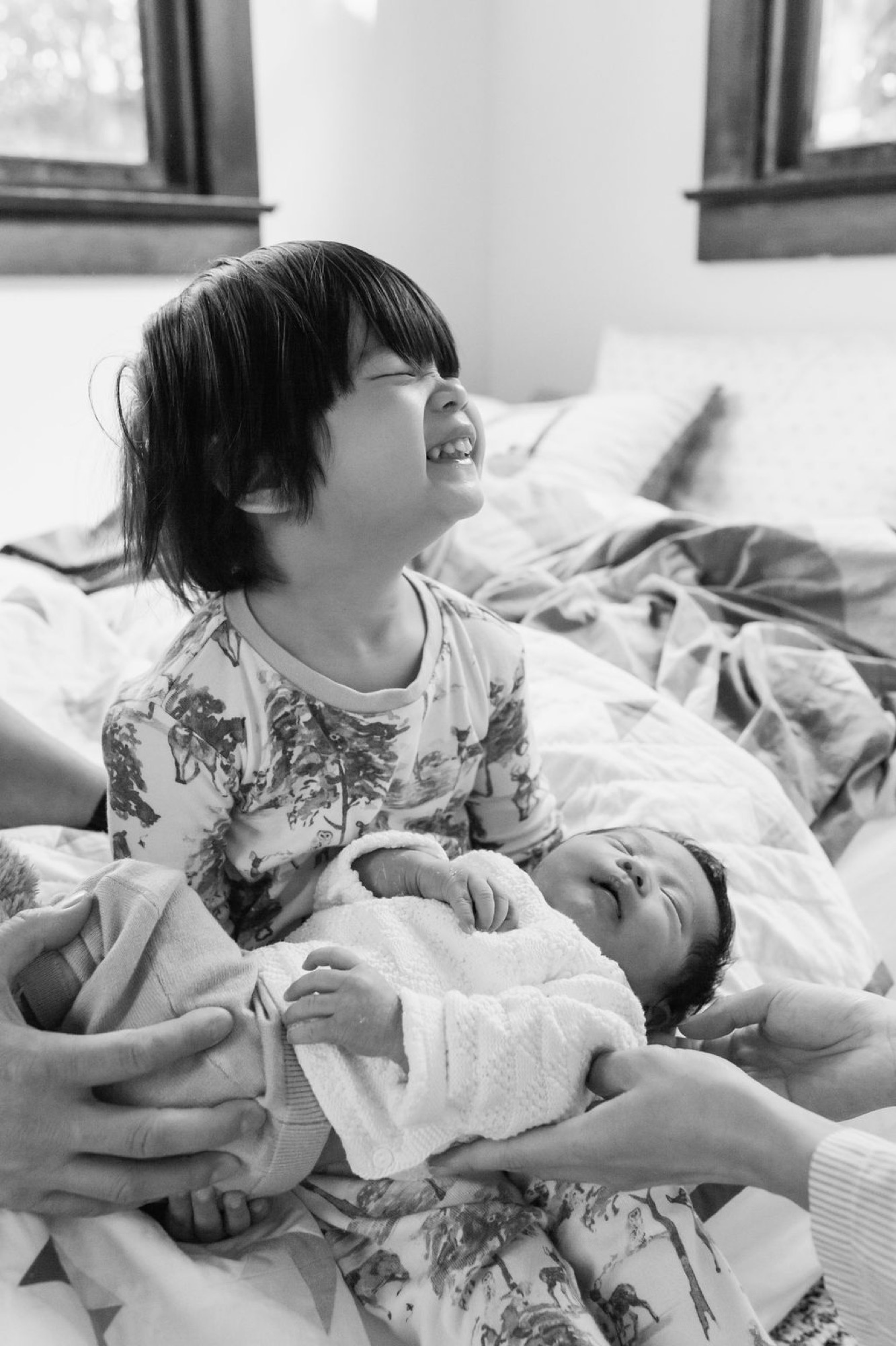 A joyful older sibling laughs with head tilted back while cradling a swaddled newborn on a bedroom bed