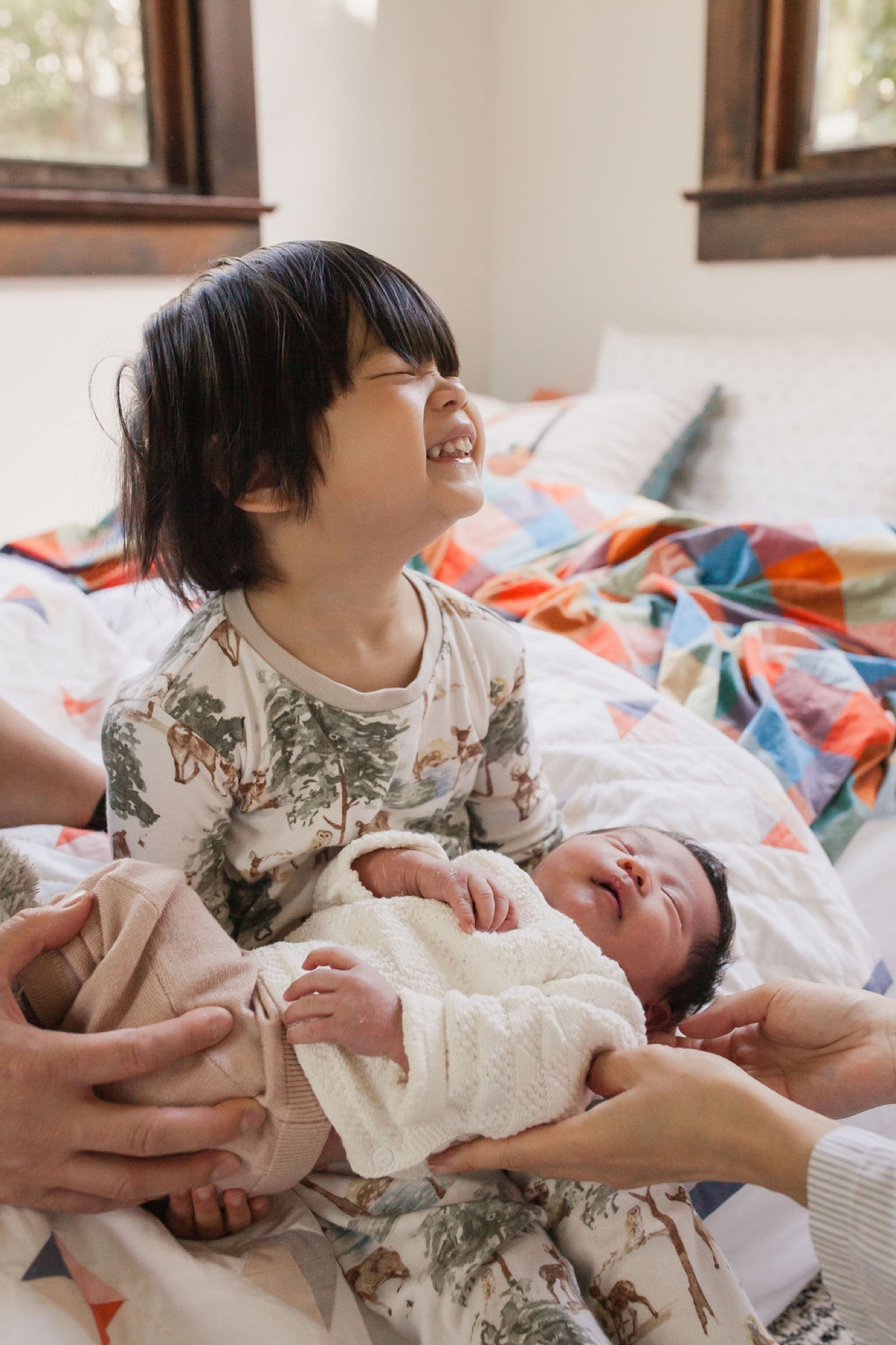 A joyful toddler in a patterned shirt holds her newborn sibling wrapped in cream knit, beaming with delight on a colorful quilt