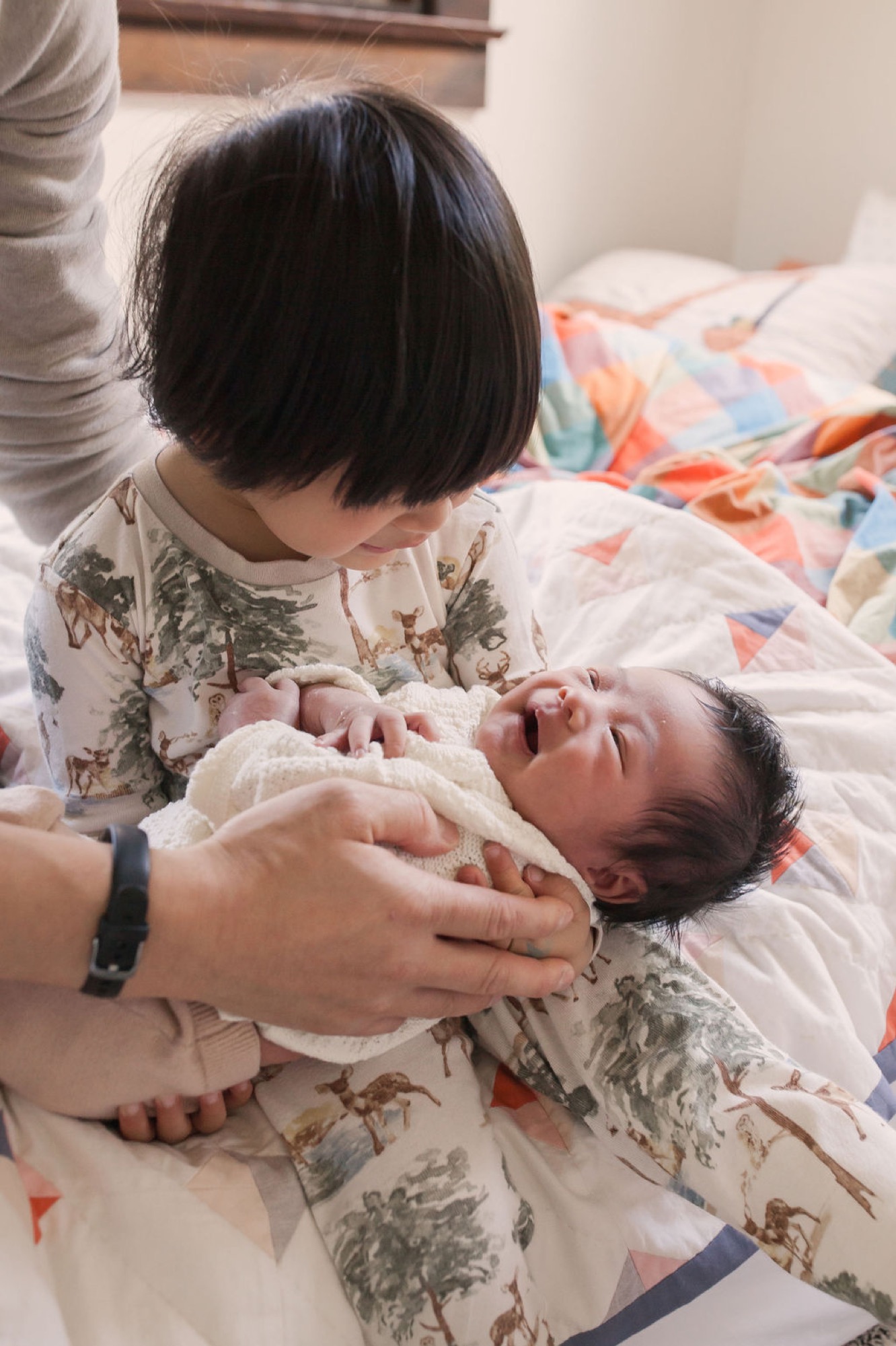 A toddler in a nature-print dress gently holds her newborn sibling wrapped in cream fabric on a colorful quilt, their tender moment softly lit by natural window light