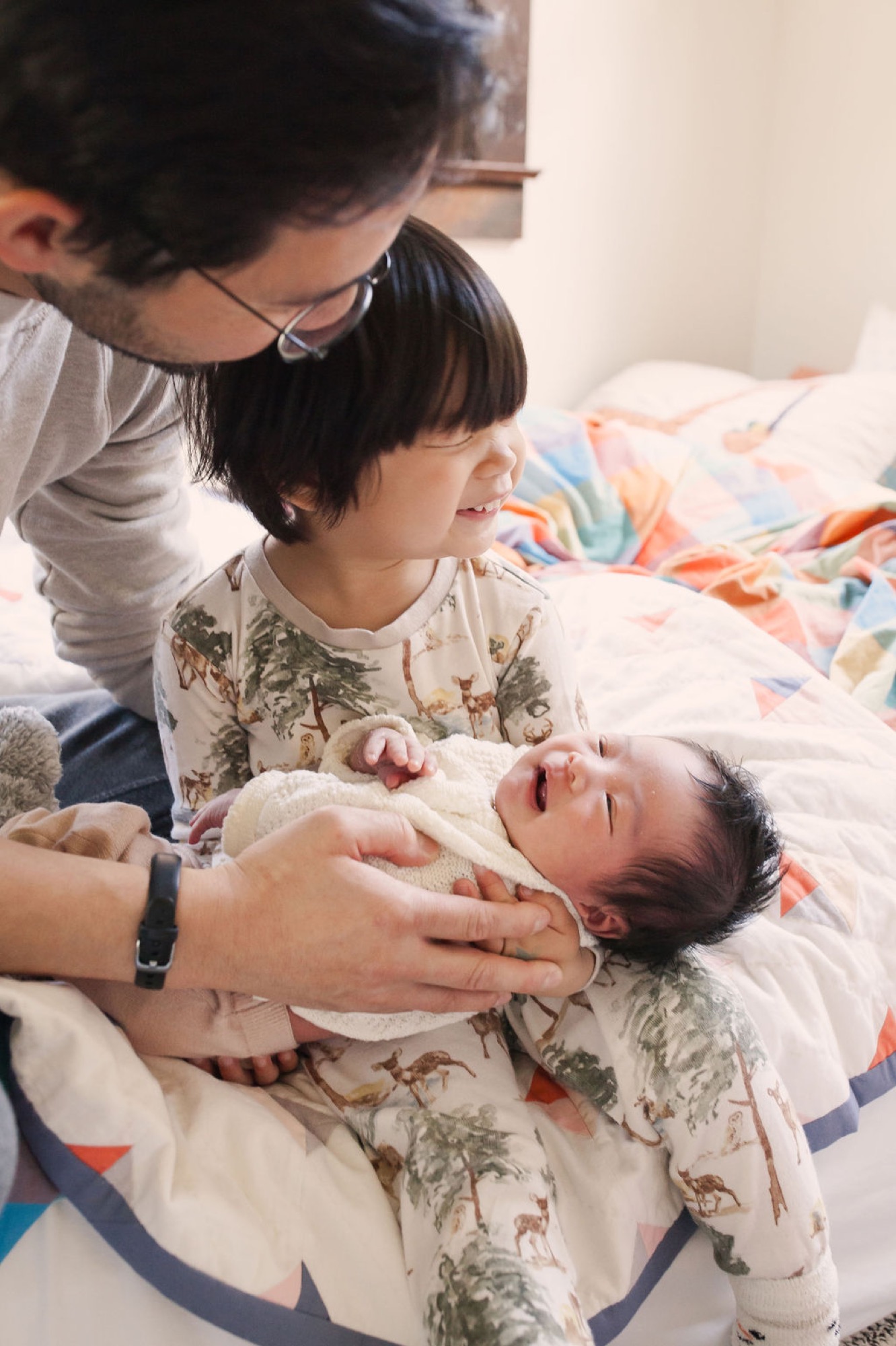 A toddler in animal-print pajamas gently holds her newborn sibling while parents lean in close, their faces lit by soft natural light filtering into the bedroom