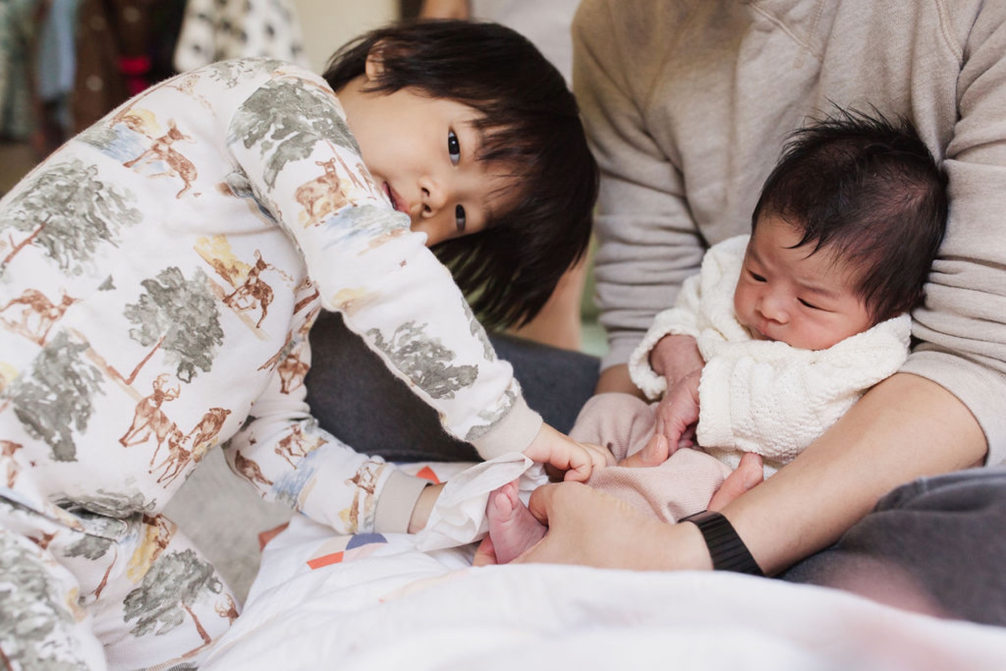 Older sibling in patterned pajamas gazes down at newborn nestled in parent's arms during intimate home session