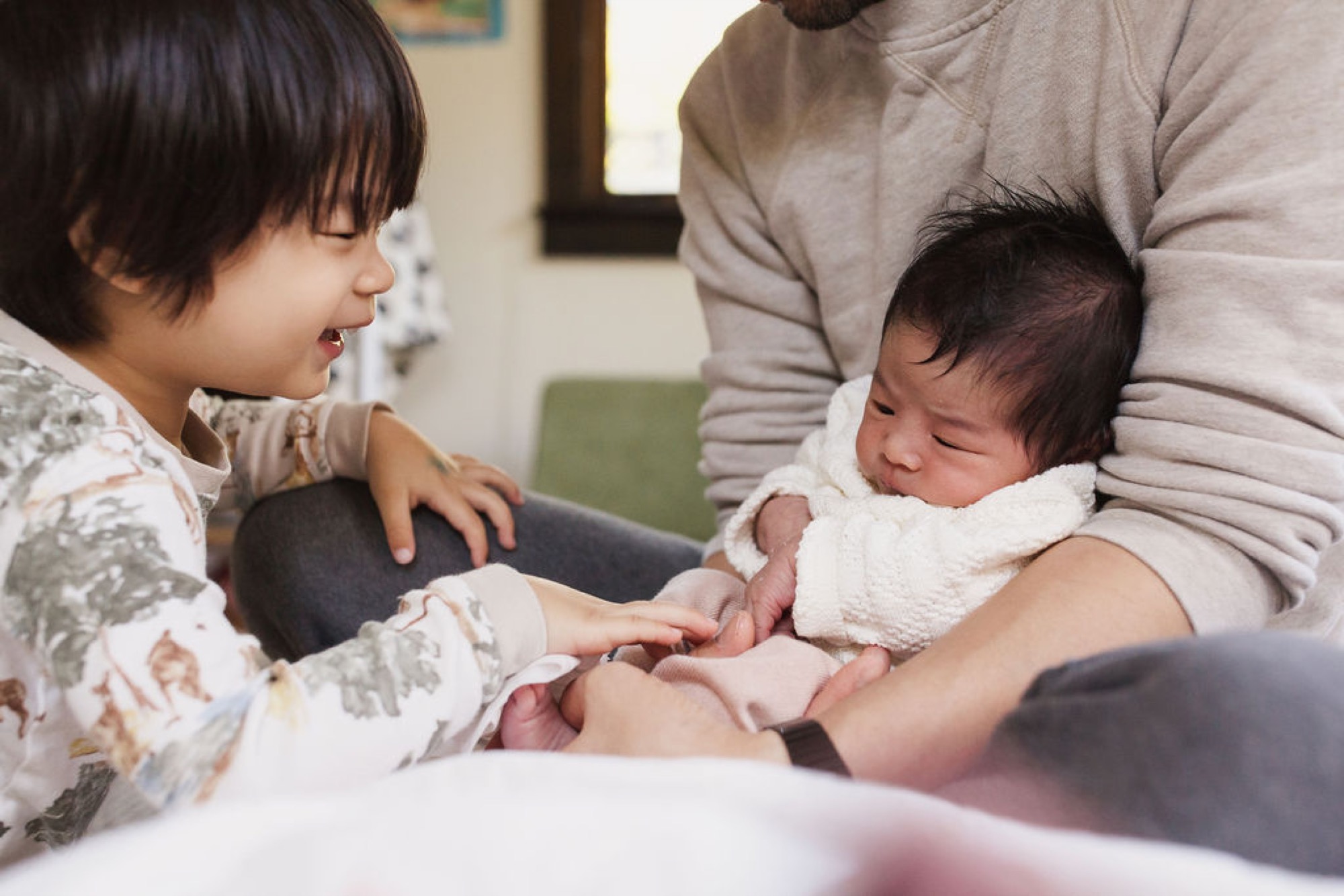 Older sibling smiles warmly while gently touching newborn's hand as father holds baby wrapped in white blanket indoors