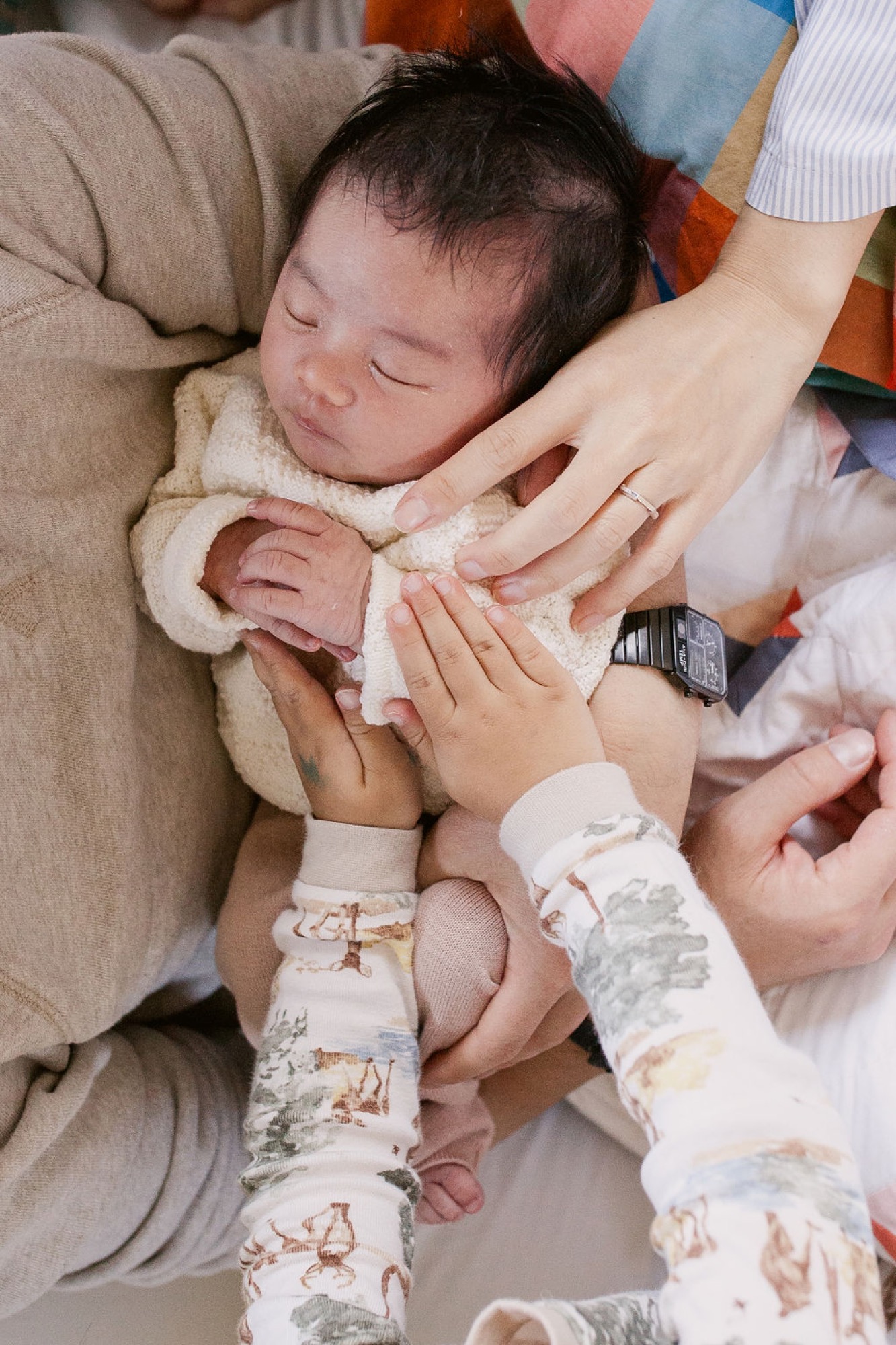 Newborn sleeps peacefully while multiple hands gently cradle and touch their tiny fingers and feet in soft indoor light