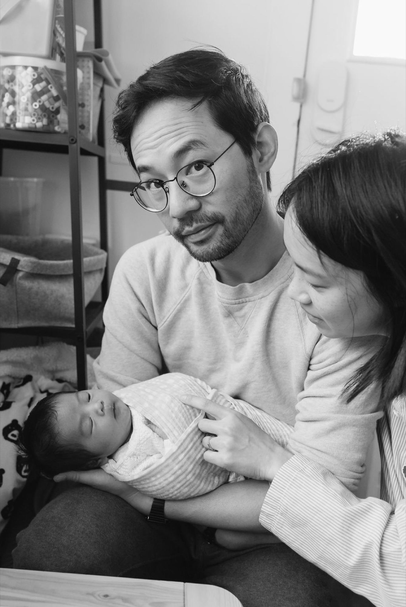A father wearing glasses gazes warmly at his newborn while the mother tenderly supports the swaddled infant in their home