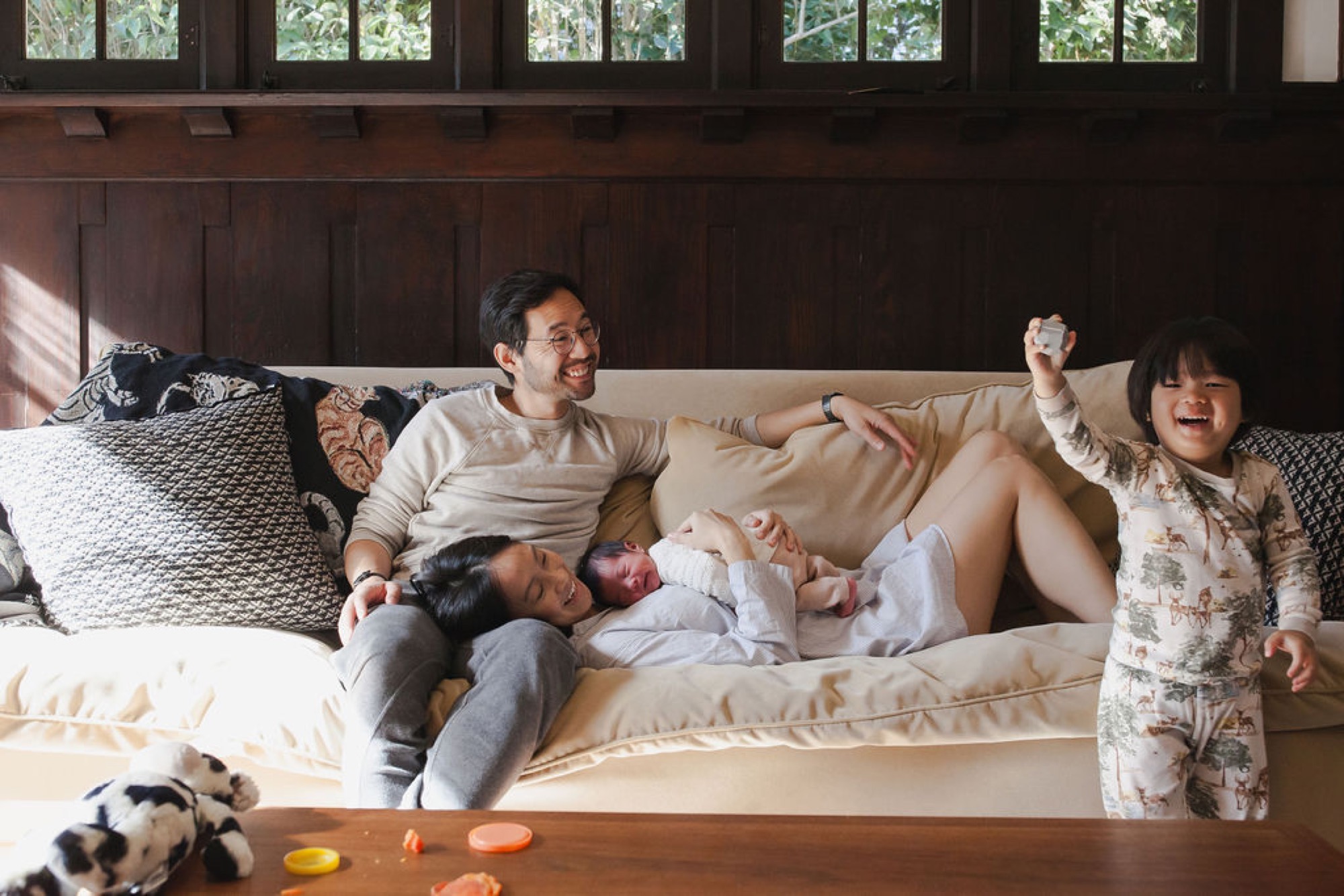 A joyful family of four lounges on a cream sofa, bathed in warm sunlight streaming through high windows, with the toddler celebrating while parents beam at their newborn
