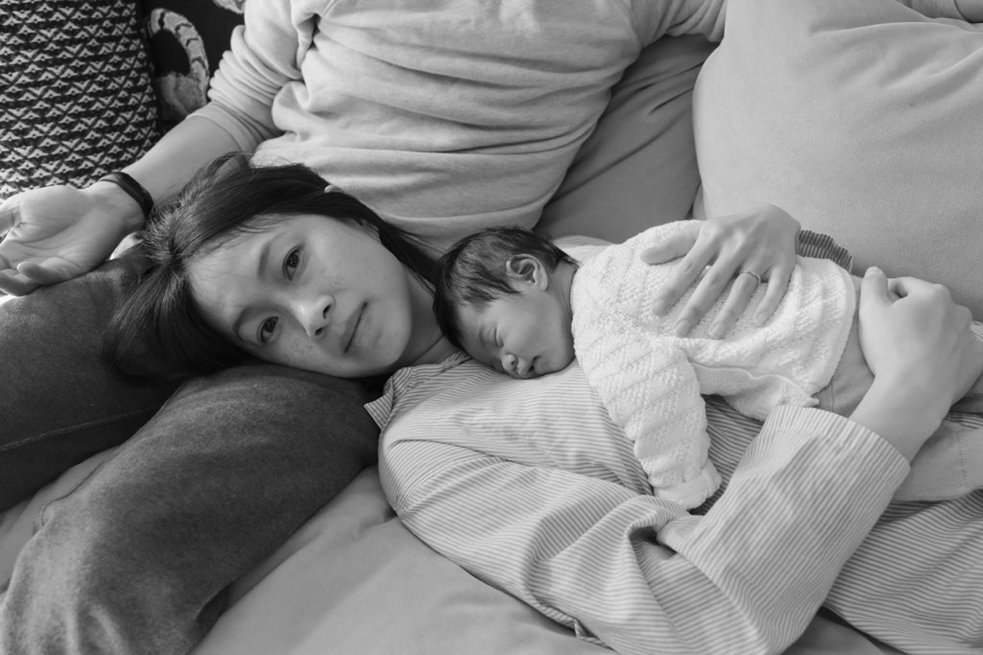 Older sister gazes at camera while cradling sleeping newborn, parents' hands tenderly framing both children on soft bedding