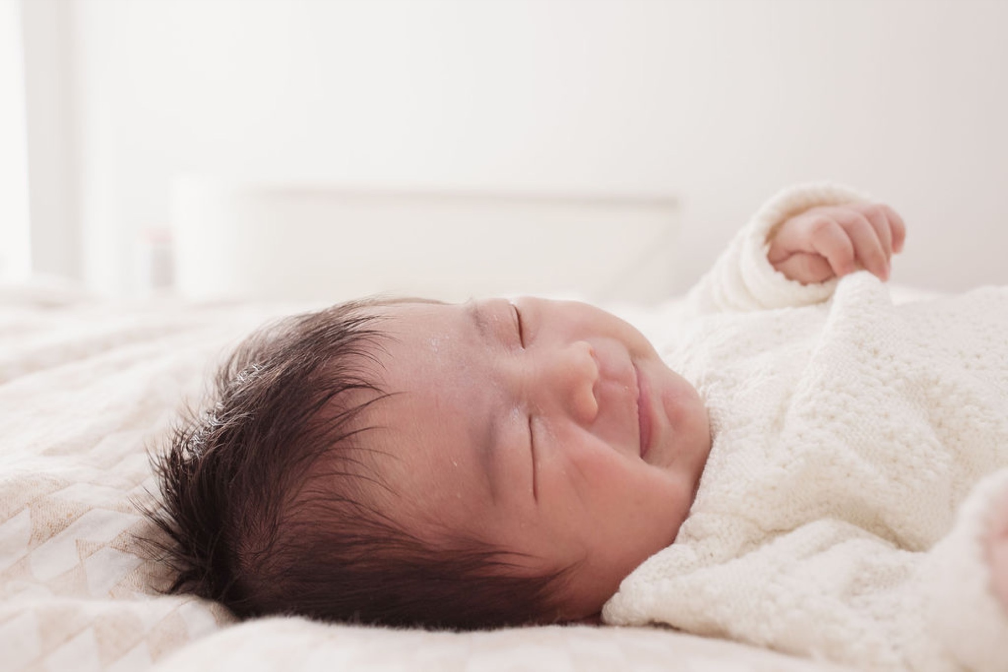 Newborn with dark hair sleeps peacefully on cream bedding, swaddled in soft white fabric with one tiny fist visible