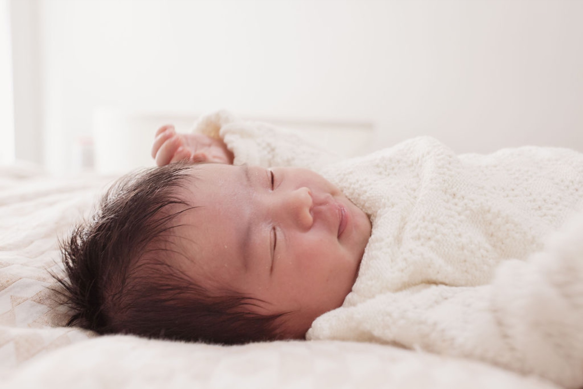 Newborn sleeps peacefully swaddled in cream blankets, dark hair visible against soft white linens in bright natural light