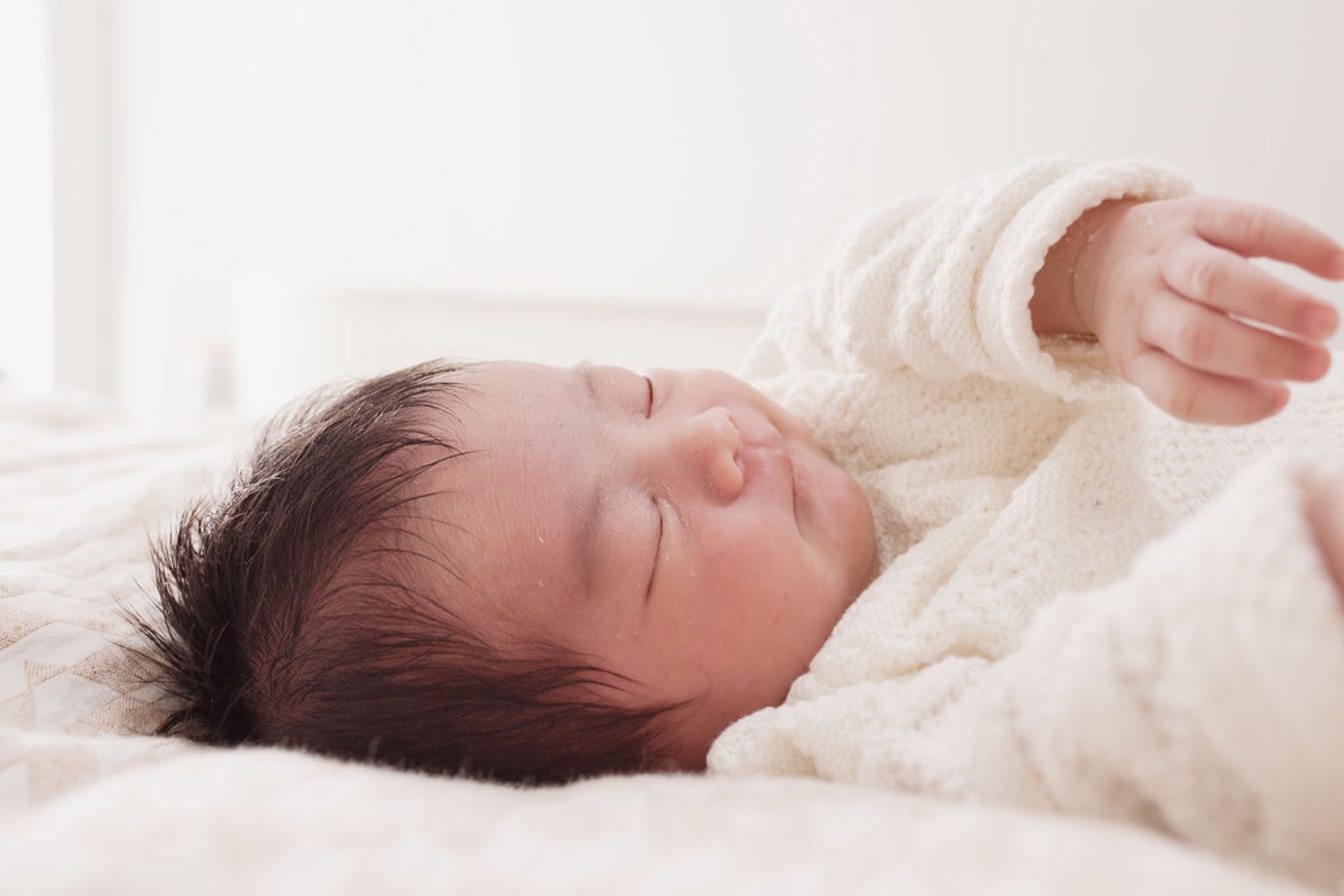 Newborn with dark hair sleeps peacefully swaddled in cream knit, tiny hand raised against soft white bedding in bright natural light