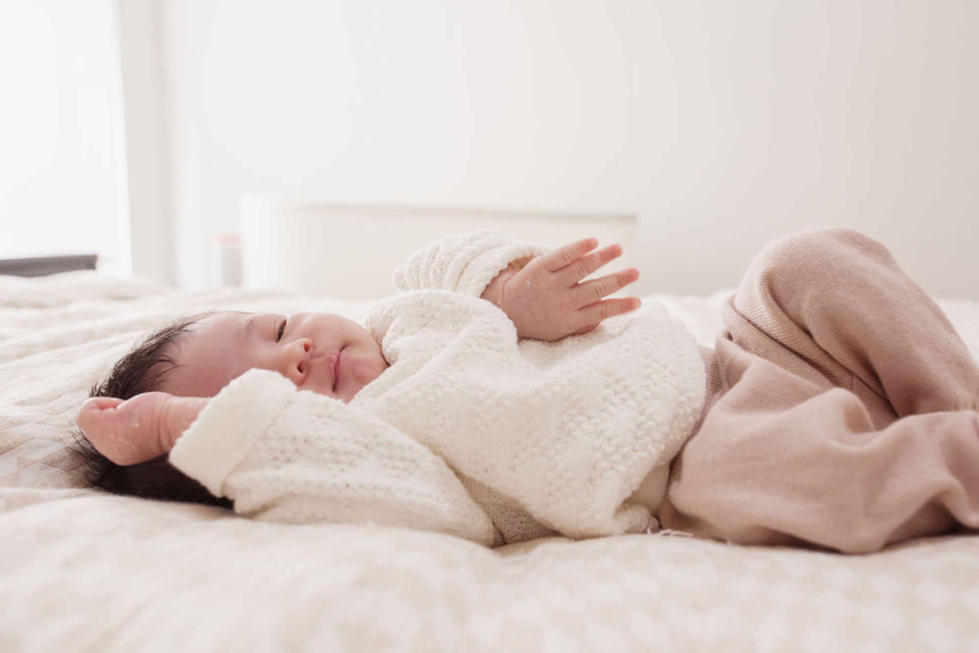 Newborn sleeps peacefully in cream knit swaddle on soft bedding, tiny hand raised in contentment, natural window light casting gentle shadows