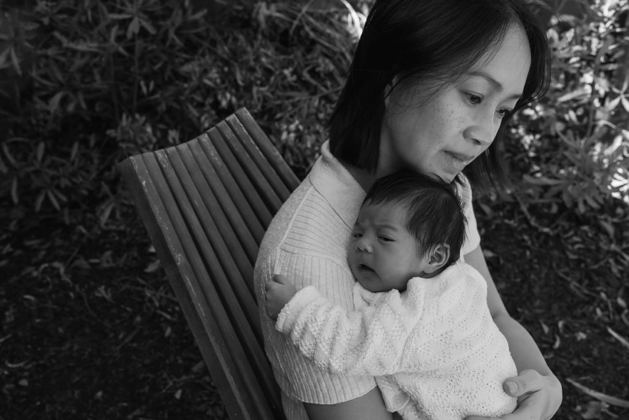 Mother gazes downward while cradling her infant in a ribbed white sweater against dense foliage backdrop