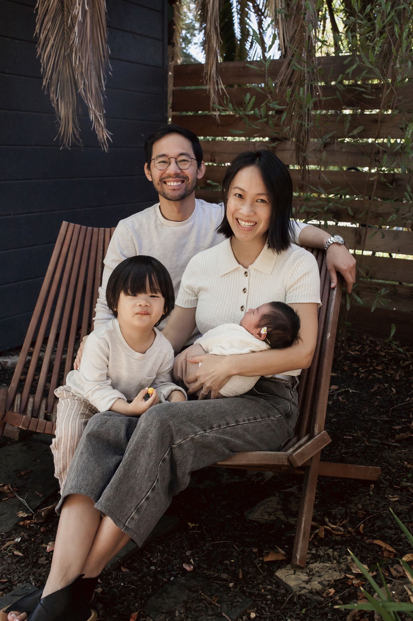 A young family of four sits together on a wooden bench surrounded by hanging dried palms, all smiling warmly at the camera in dappled afternoon light