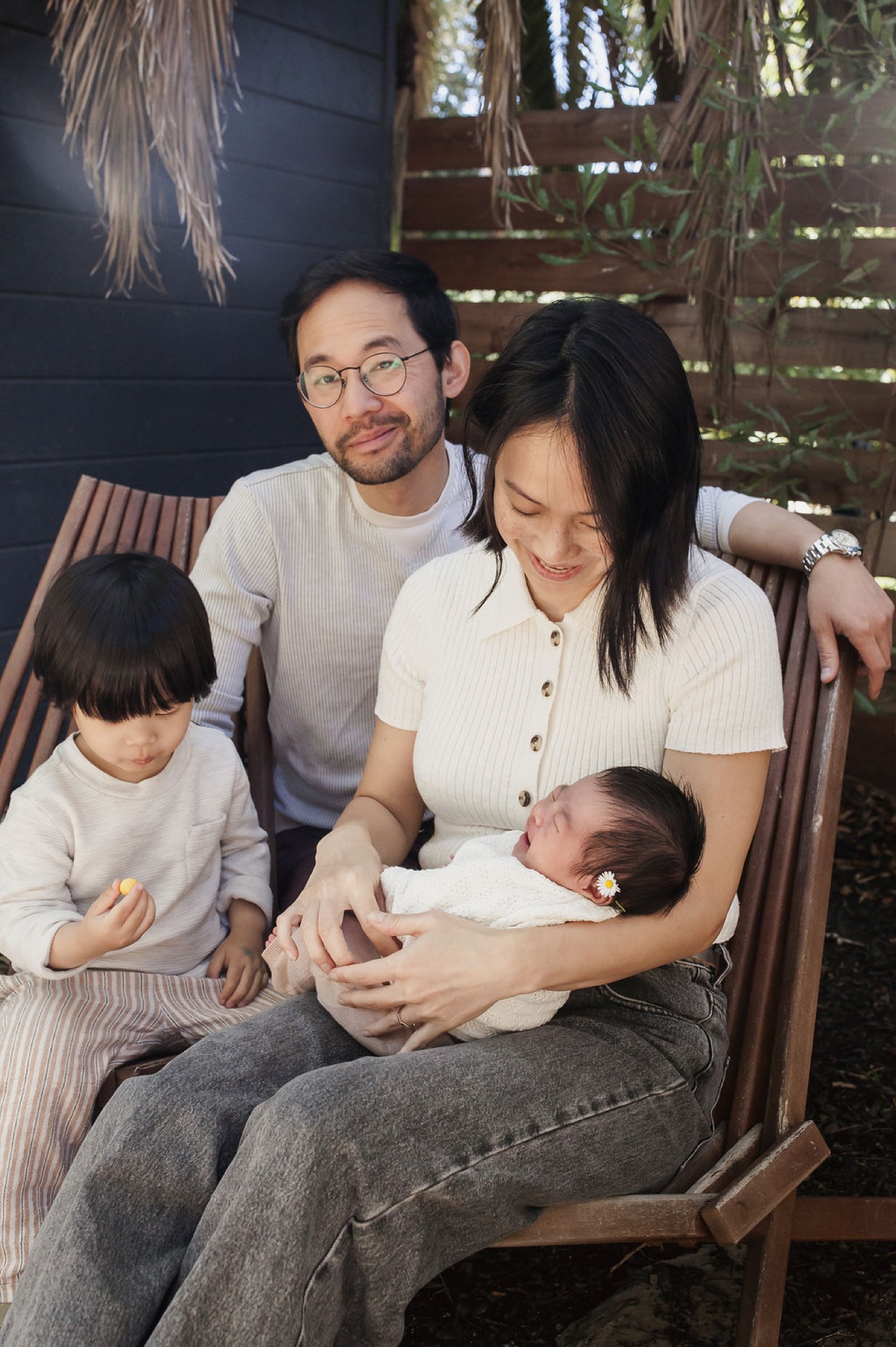 A family of four sits together on a wooden bench beneath hanging vines, the mother cradling a newborn while her older daughter and father look on warmly in soft, dappled light