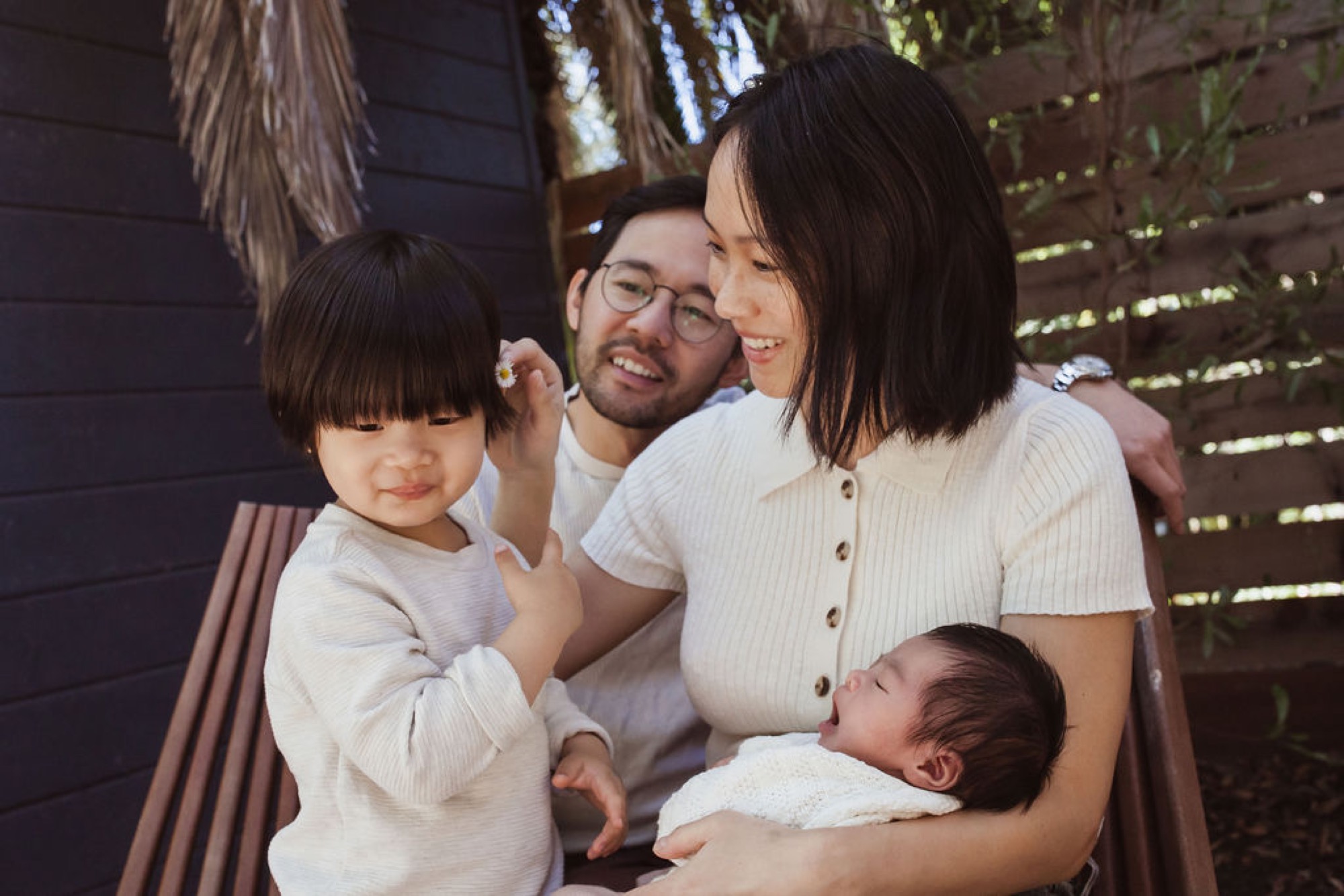 Young family shares genuine laughter together in dappled garden light, mother holding newborn while father embraces toddler with warm affection